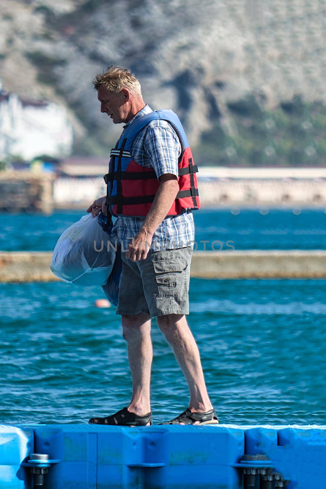 Elderly man in a life jacket goes on the dock at sea. View from the side. Sudak, Crimea - 4 October, 2020 by Essffes