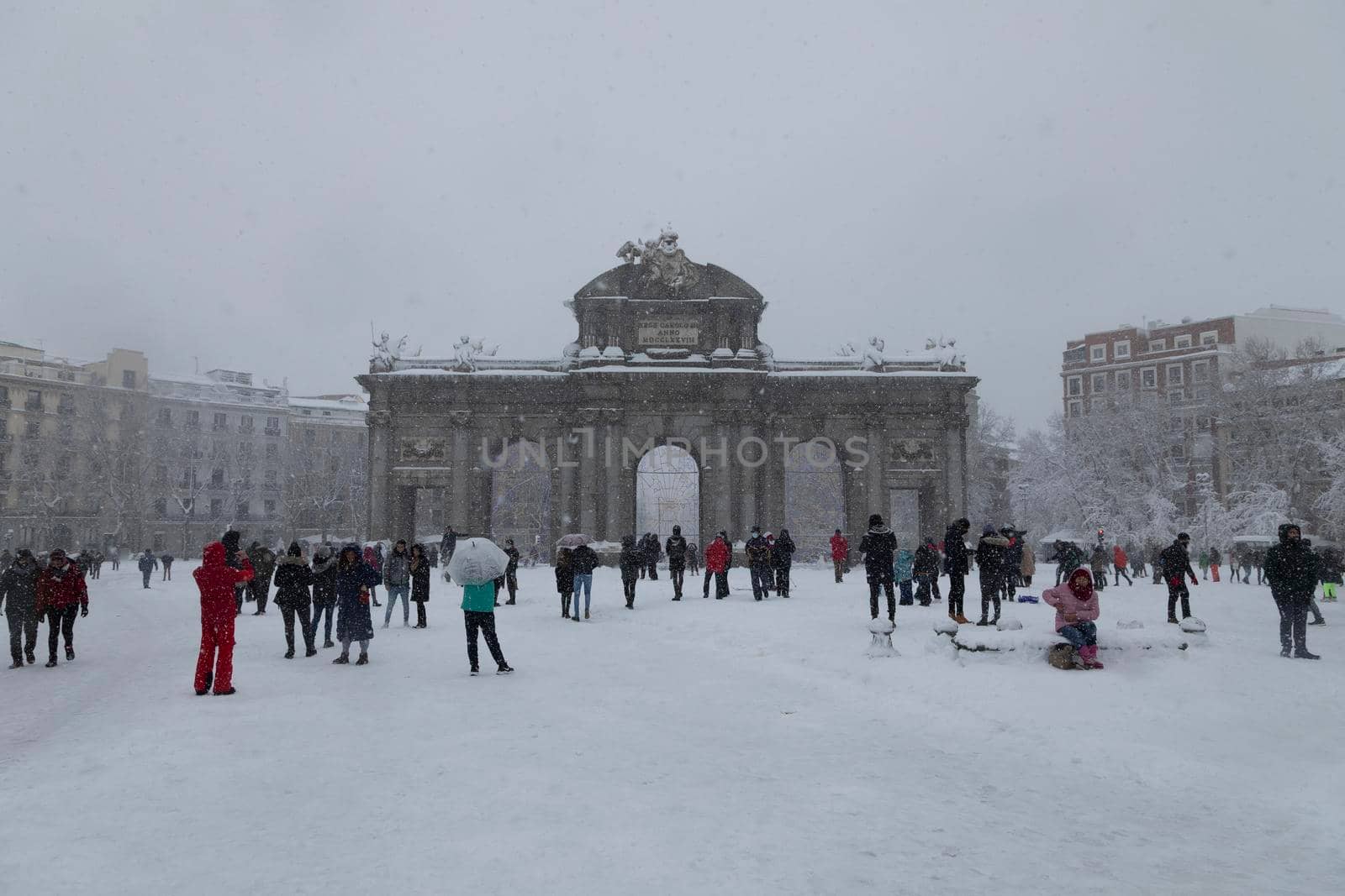 Madrid, Spain - January 09, 2021: The famous Alcala gate, frozen and covered with snow, surrounded by people walking, on a snowy day, due to the polar cold front Filomena.