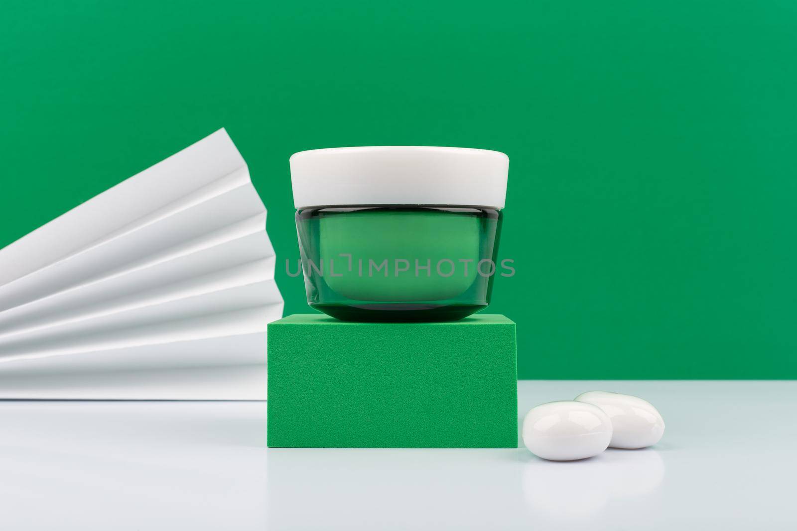Still life with green cream jar on green podium on white glossy table against green decorated with waver background. Concept of luxurious organic skincare for smooth, firm and young looking skin