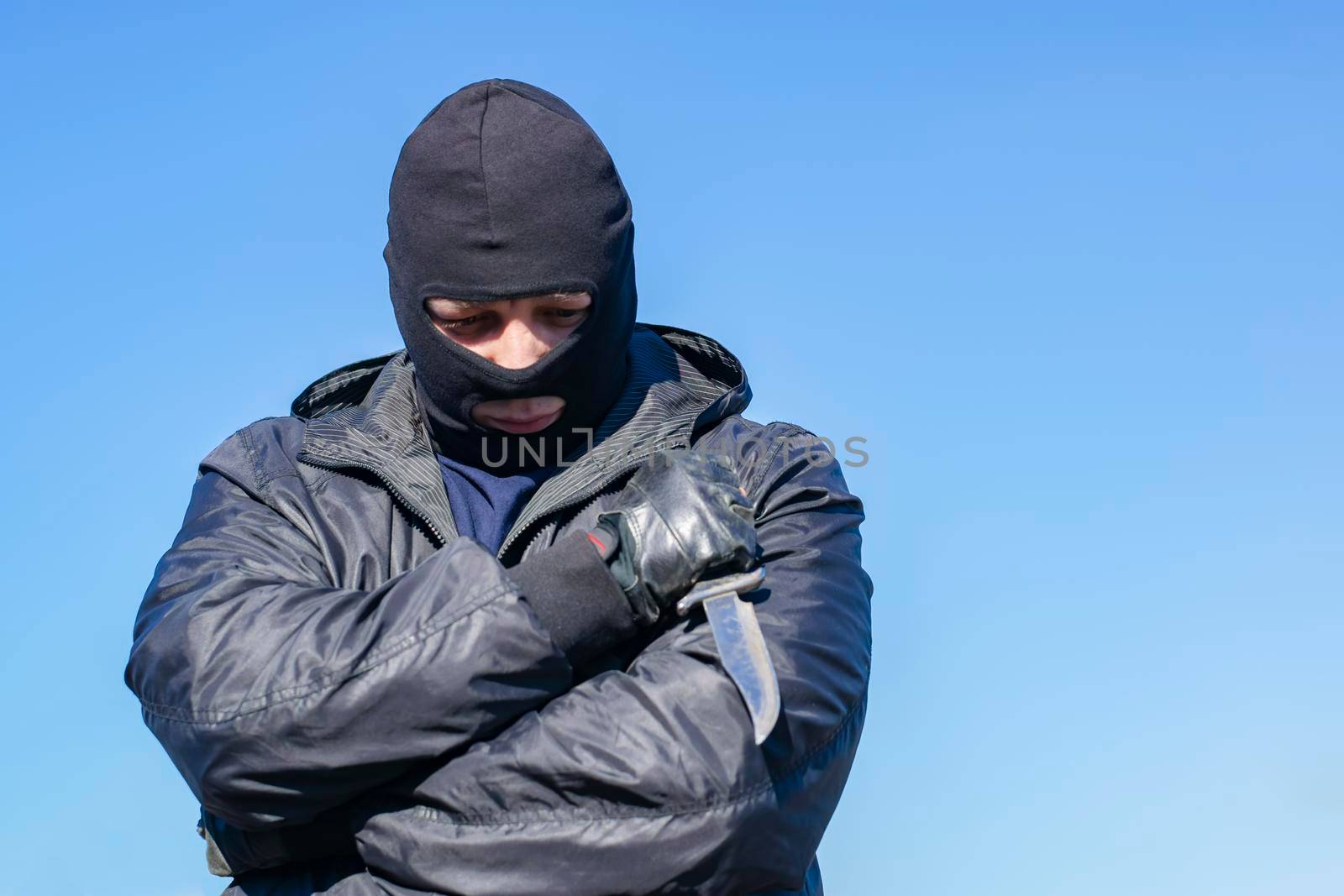 a sad, thoughtful bandit stands in a black mask and with a knife against the blue sky