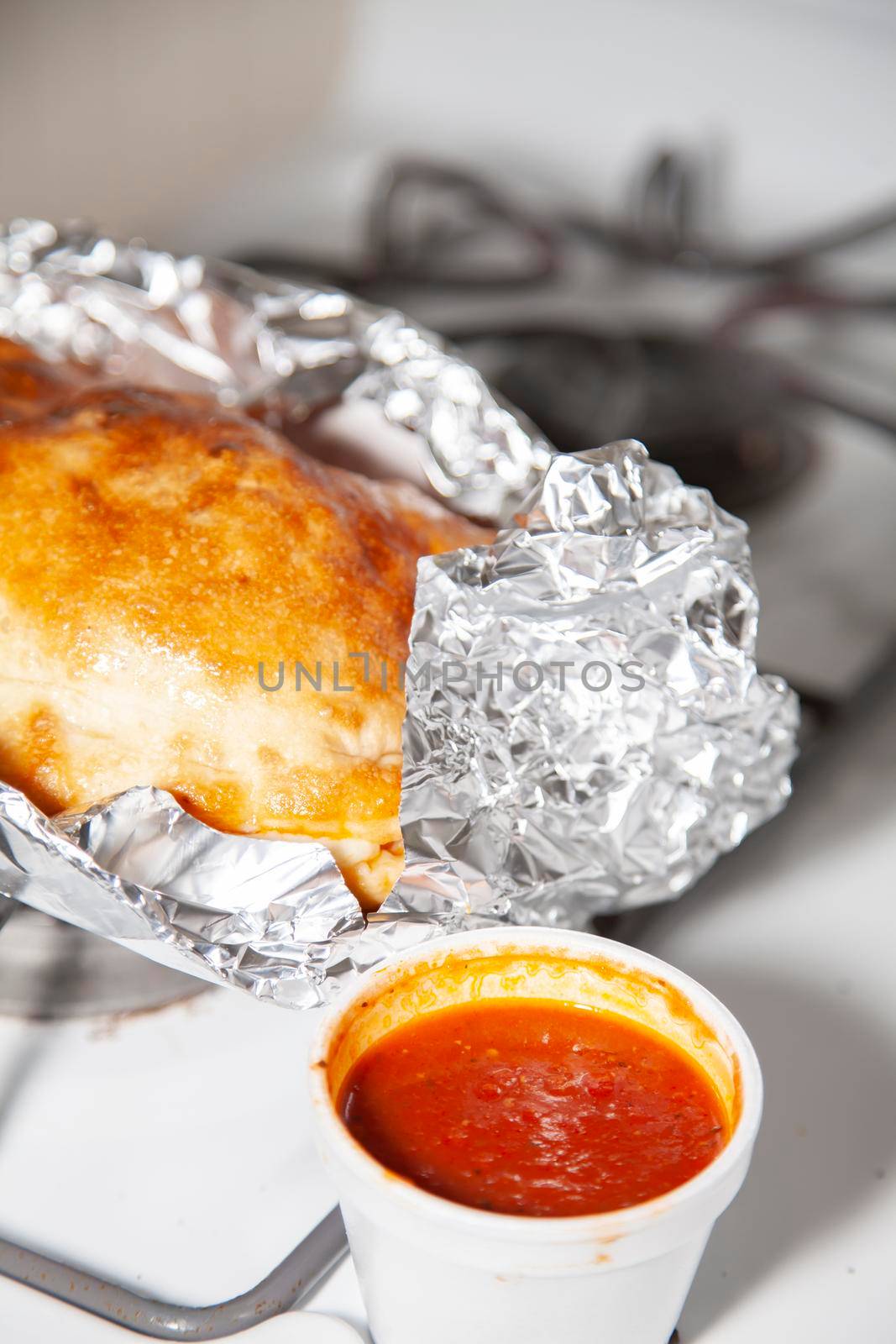 Glazed outter bread of a fresh calzone on foil next to a styrofoam cup of marinara sauce
