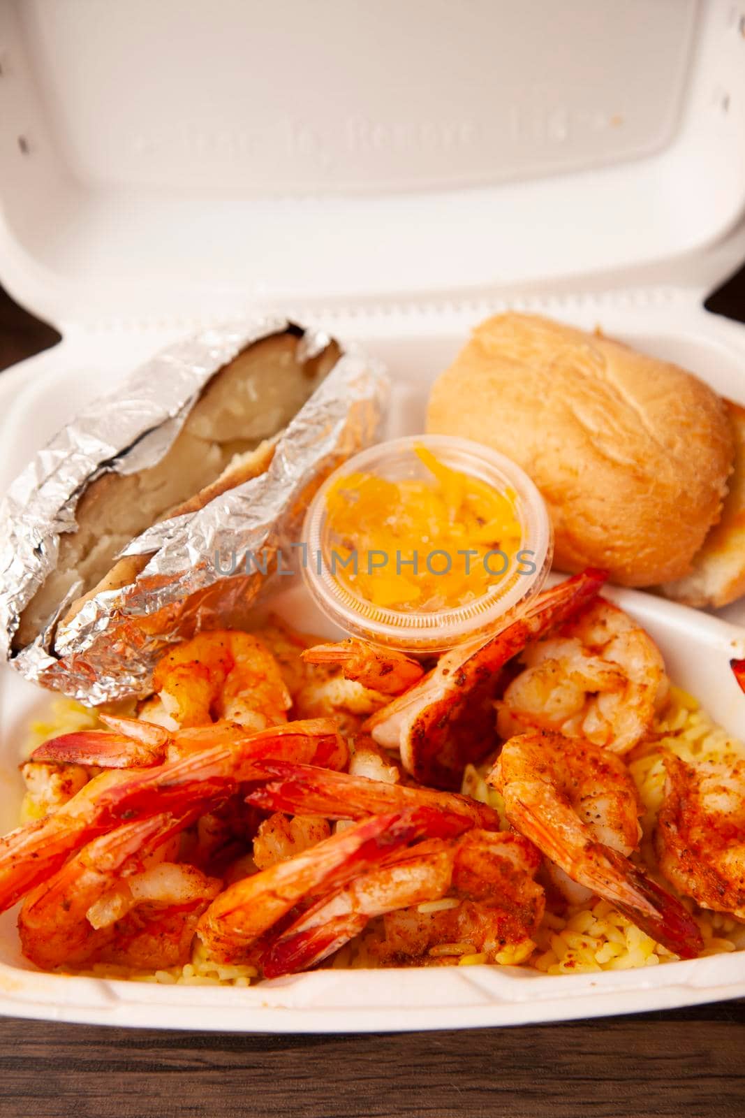Grilled shrimp, baked potato, and roll with a container of sauce in a white, styrofoam carryout box