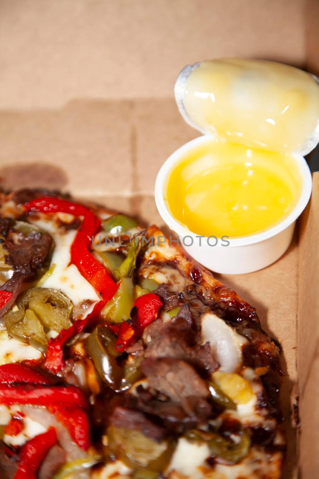 Steak, red bell pepper, jalapeño, green bell pepper, and onion pizza in a cardboard carry out container with garlic dipping sauce