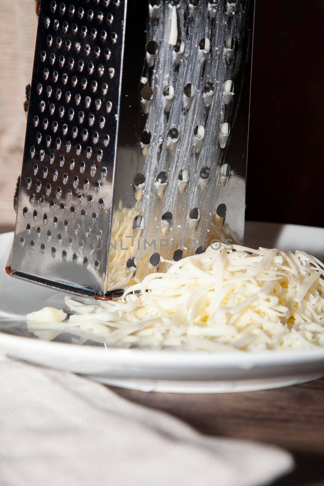 Pile of shredded white cheese next to a large, metal cheese grater on a white plate next to a tan napkin on a wooden table
