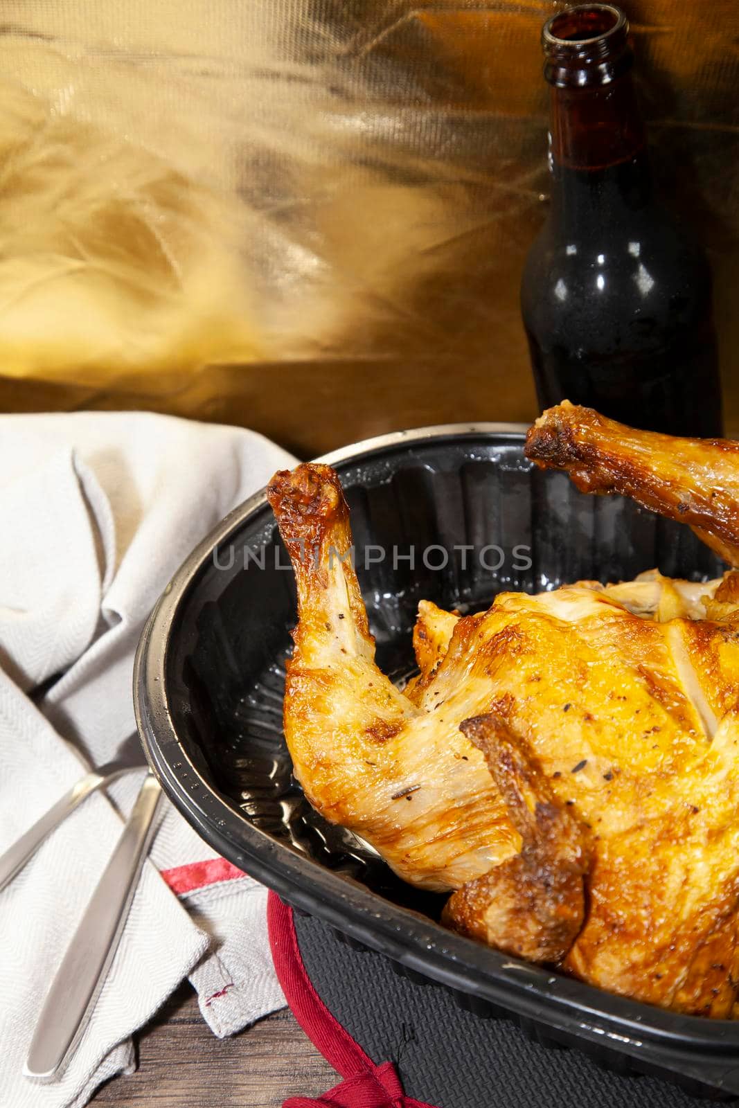 Whole roasted chicken in a black carryout container and a bottle of beer next to a grey napkin and silverware