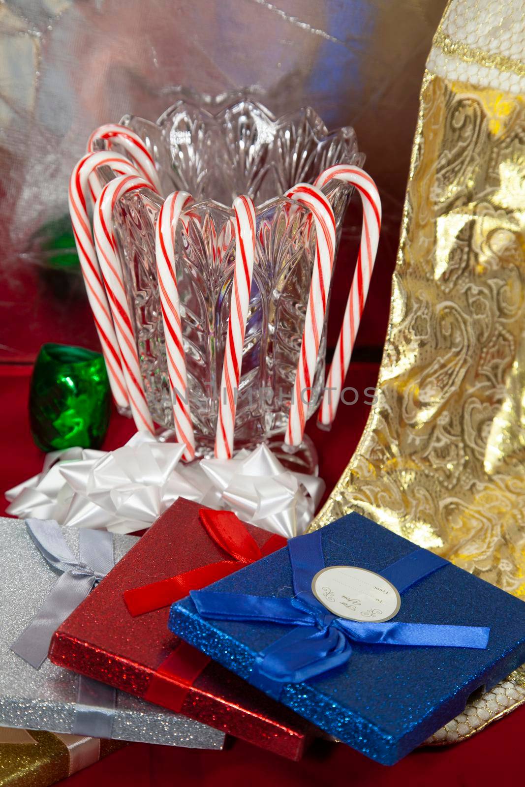 Red striped candy canes hanging on a crystal vase next to a golden stocking and silver, red, and blue presents, on a red tabletop