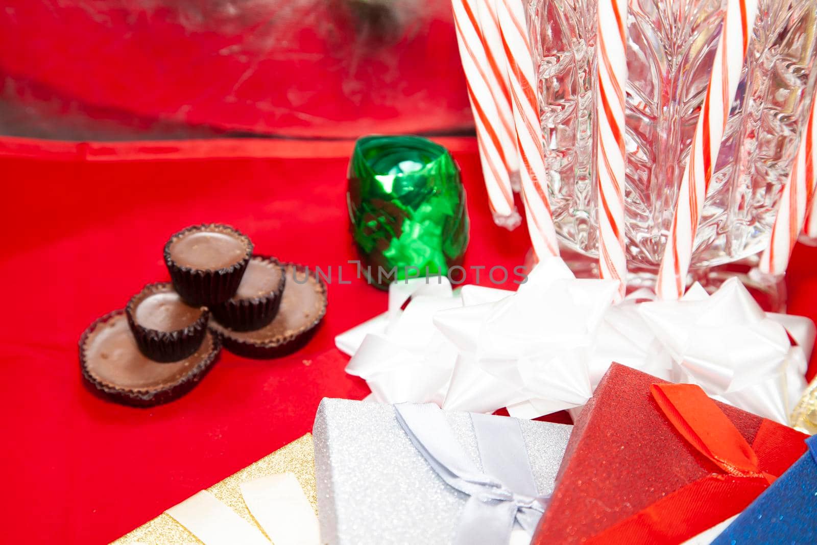Candy canes on a glass vase with chocolates, green ribbon paper and gold, silver, and red presents on a red tabletop
