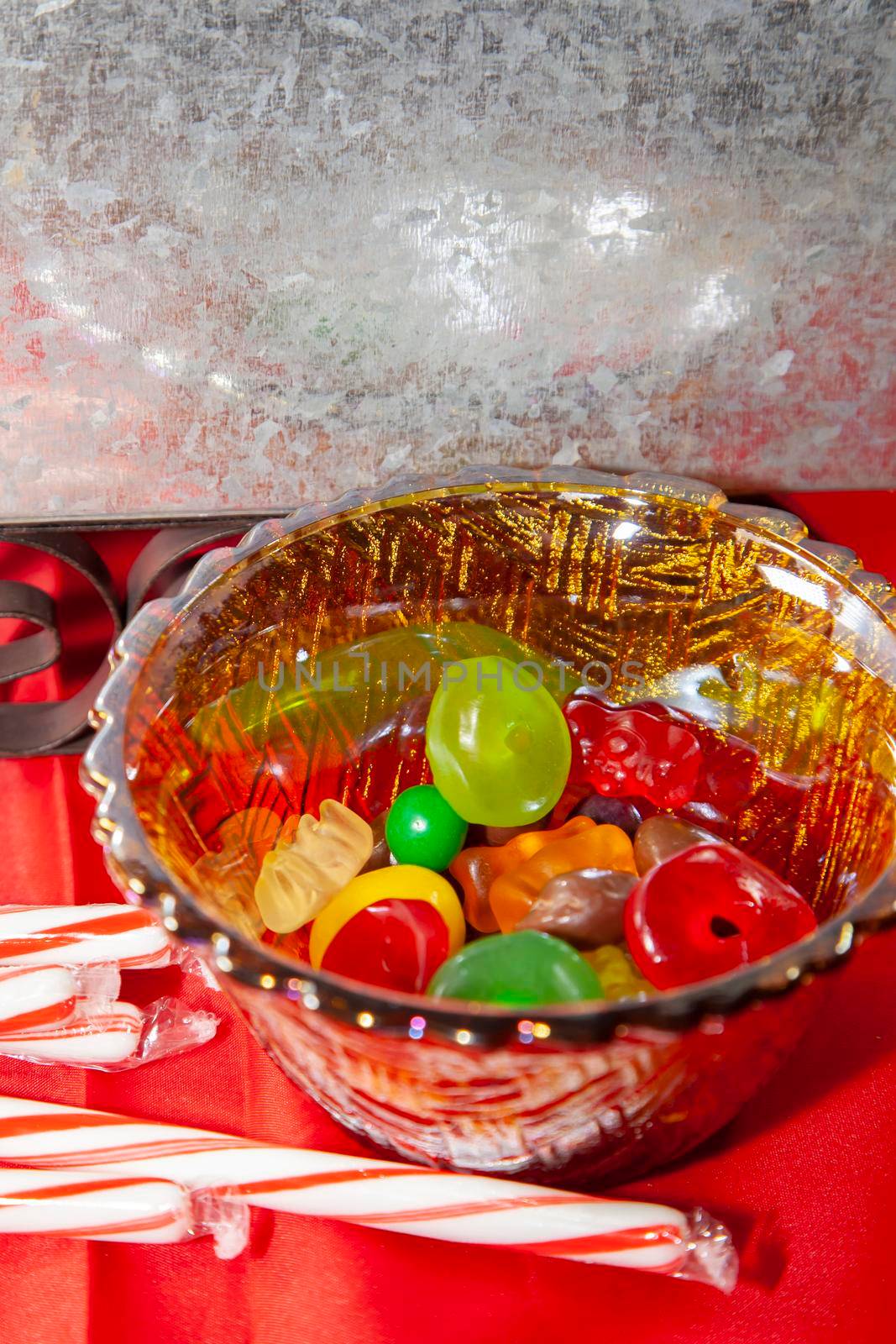 Multicolored candies in a small bowl next to candy canes on a red tabletop with a tin sleigh in the background