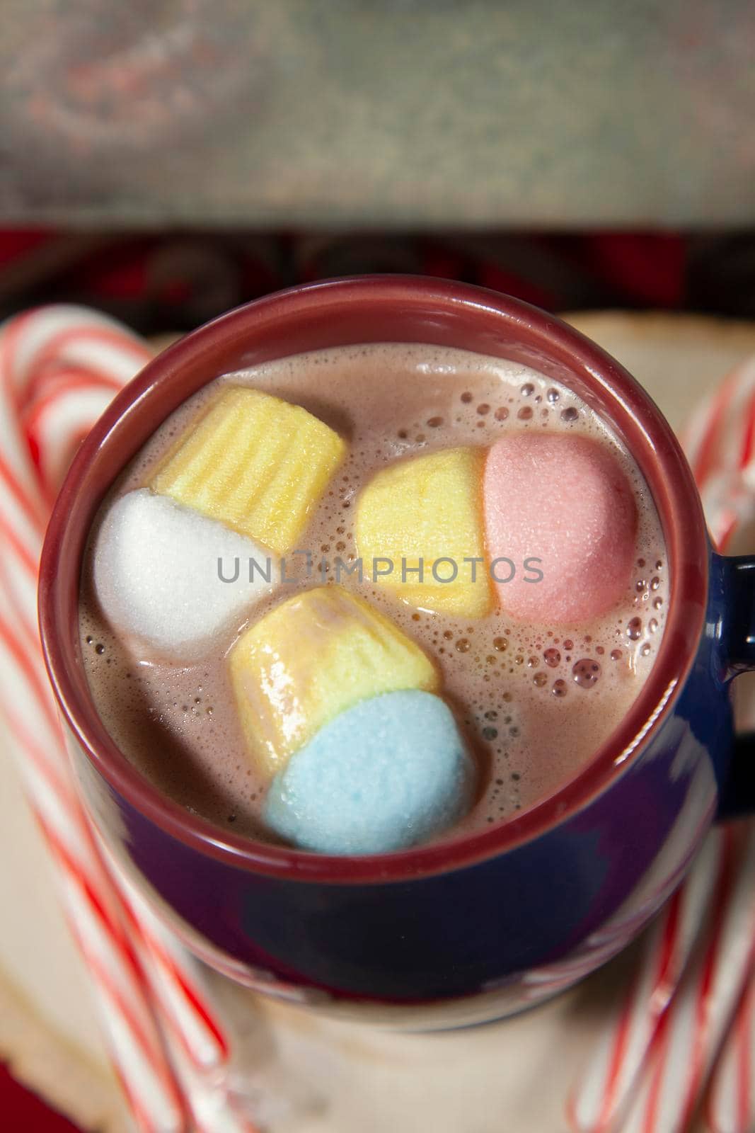 Blue and red mug with multicolored marshmallows on a wooden surface with candy canes, next to a tin background