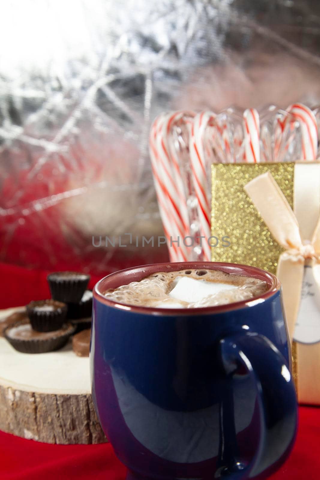 Blue and red mug filled with hot chocolate and two slightly melted marshmallows, with chocolates on a wooden stump, candy canes, and a golden present in the background