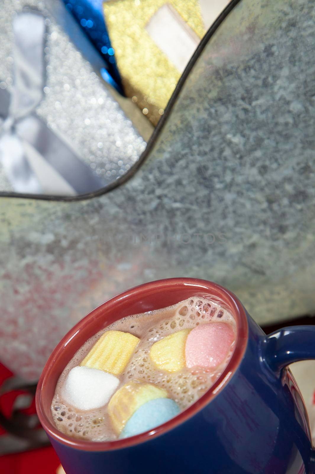 Blue and red mug with multicolored marshmallows next to a tin sleigh filled iwth silver, blue, and gold presents in the background