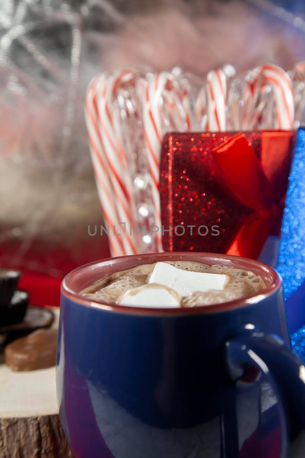 Blue and red mug filled with hot chocolate and two marshmallows, with chocolates on a wooden stump and candy canes and red and blue presents in the background