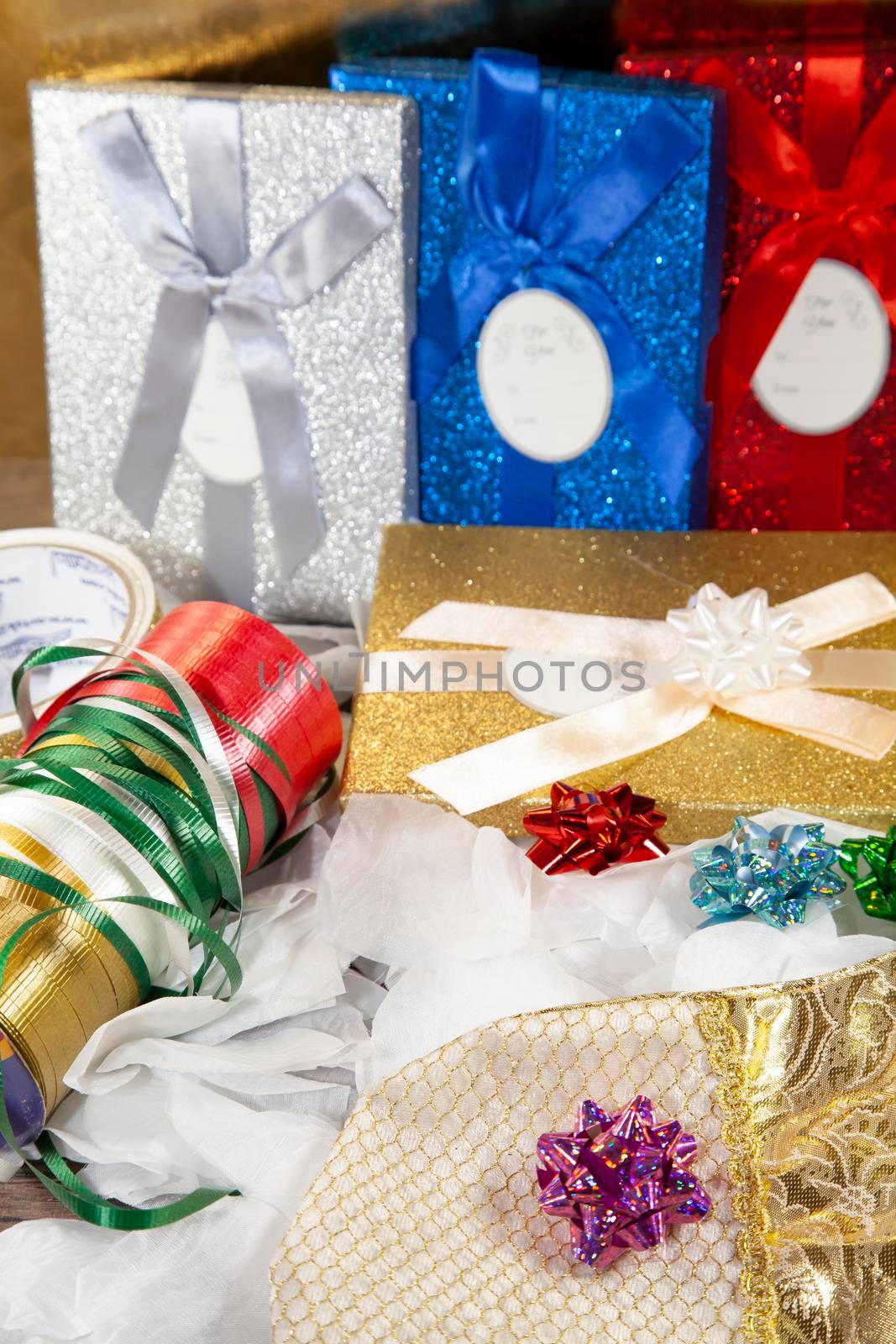 Golden gift box topped with a white bow in front of silver, blue, and red gift boxes and next to wrapping ribbon, red, blue, and green bows, and a golden stocking topped with a pinkish/purple bow