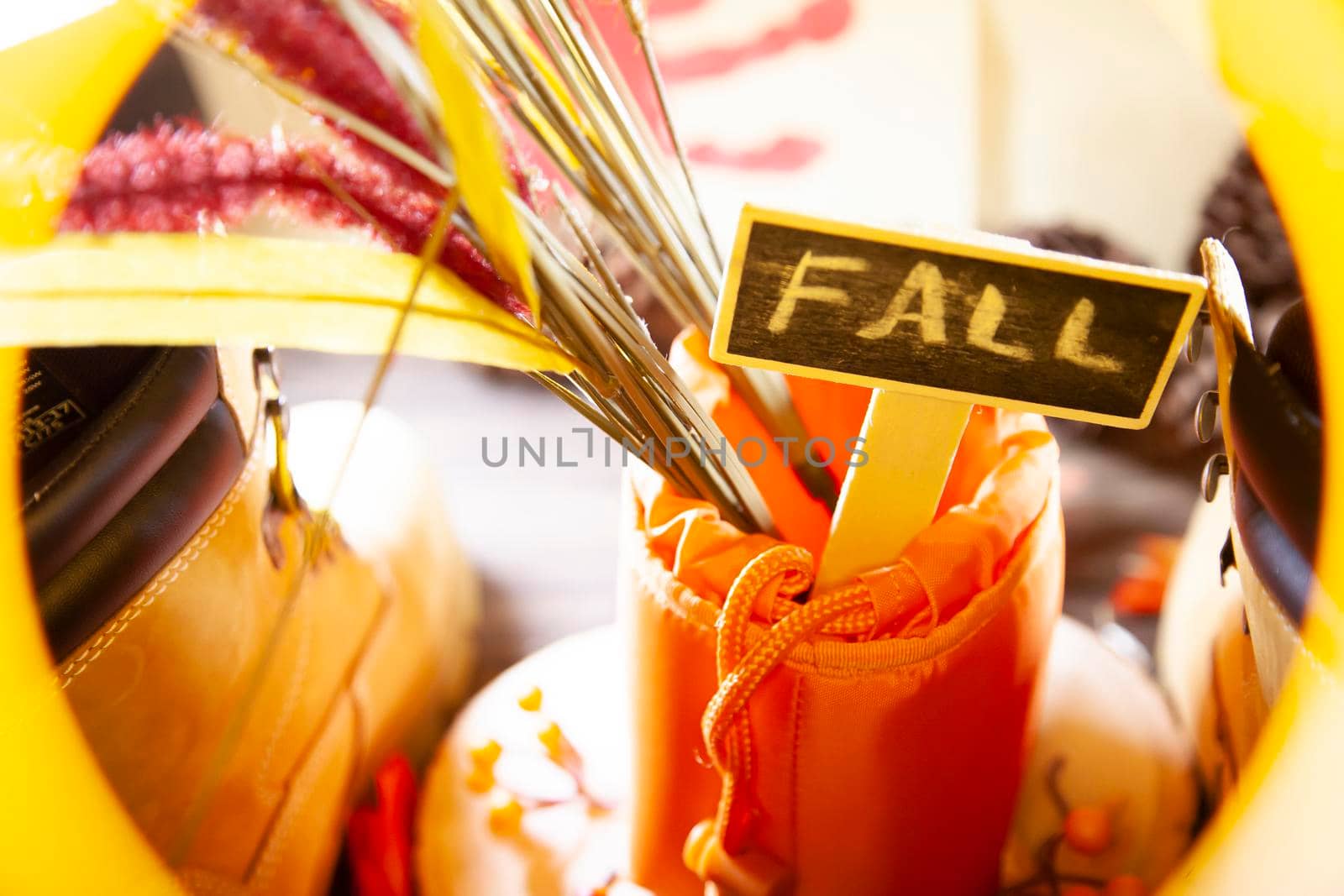 A "FALL" sign in a hunter orange pouch on a wooden slab next to orange and red berries and leaves between boots with a red hand turkey in the background framed in yellow