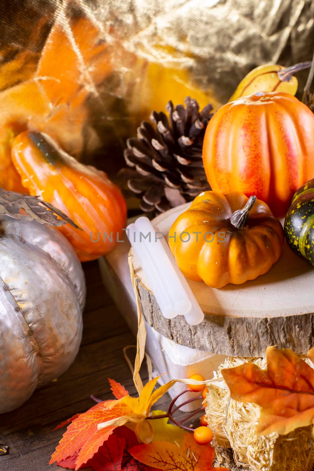 Small orange pumpkins, orange squash, pinecones, a green squash, and glue sticks for crafting on wood stacked on leaf-covered hay next to a silver pumpkin with a golden background
