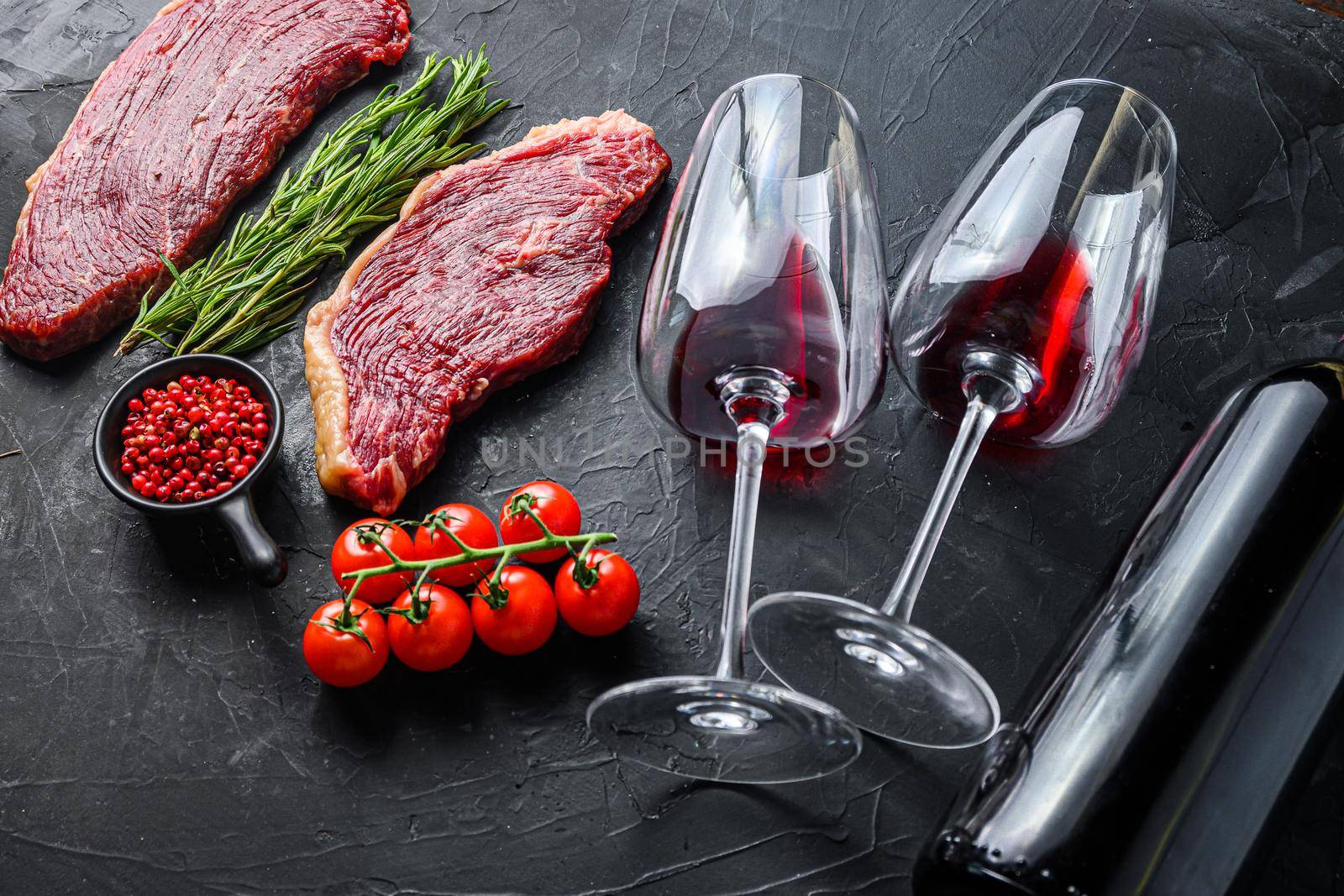 Raw picanha steaks with seasonings and herbs near bottle and glass of red wine, over black textured background side view