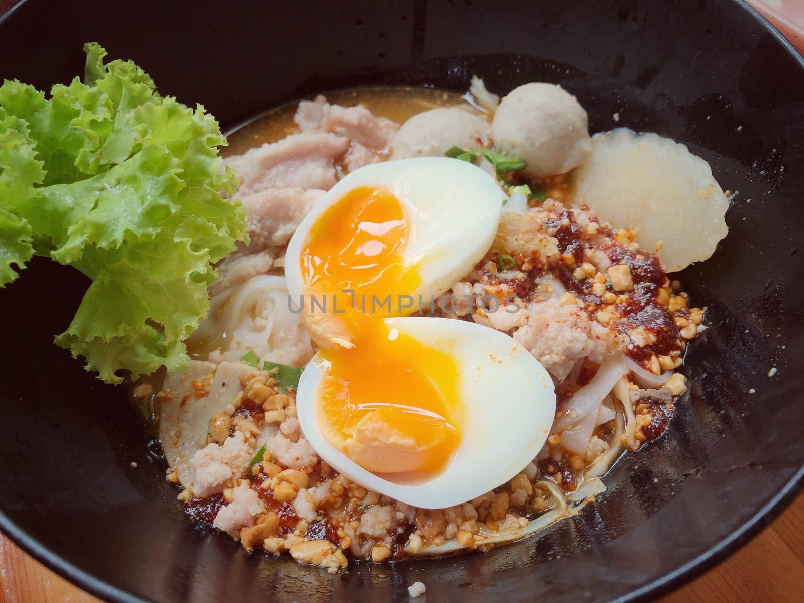 Thai spicy pork noodle soup with coconut milk, vegetables herbs and onsen tamago eggs  in black bowl with sesame seeds and peanut on wooden table, view from above by NarinNonthamand
