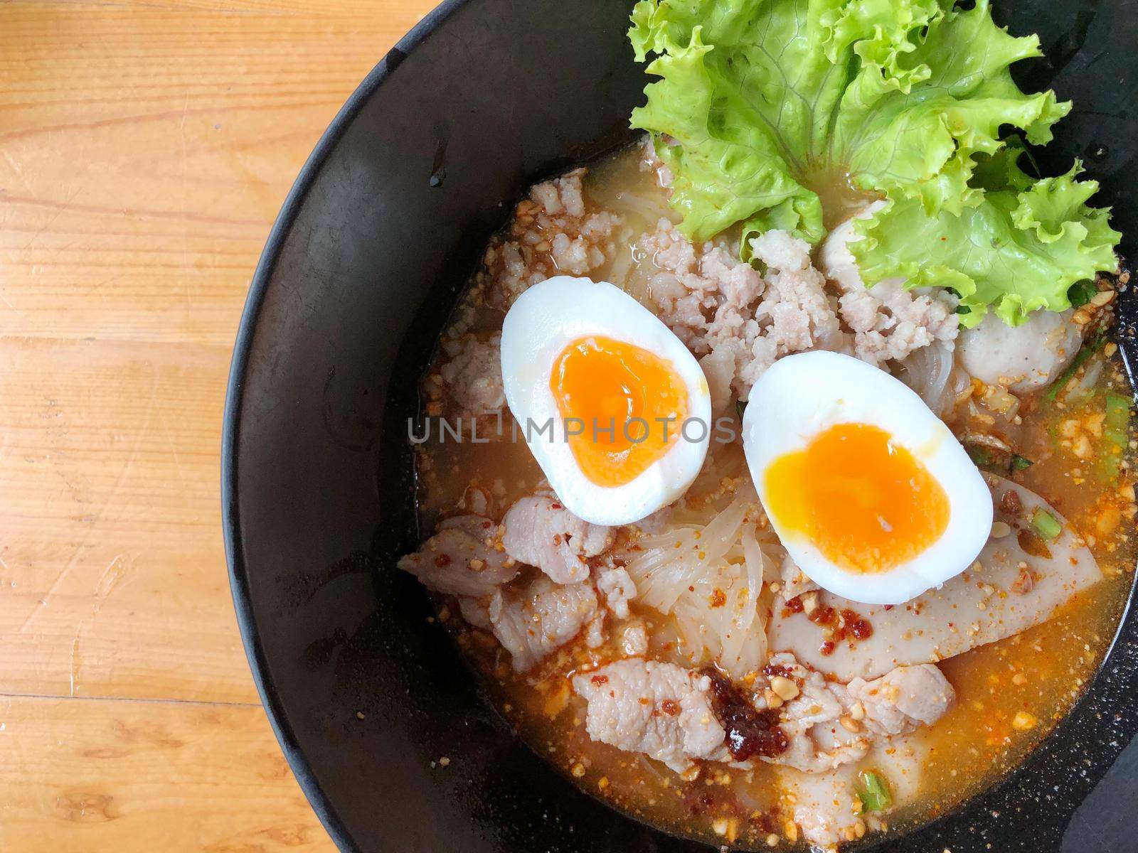 Thai spicy pork noodle soup with coconut milk, vegetables herbs and onsen tamago eggs  in black bowl with sesame seeds and peanut on wooden table, view from above