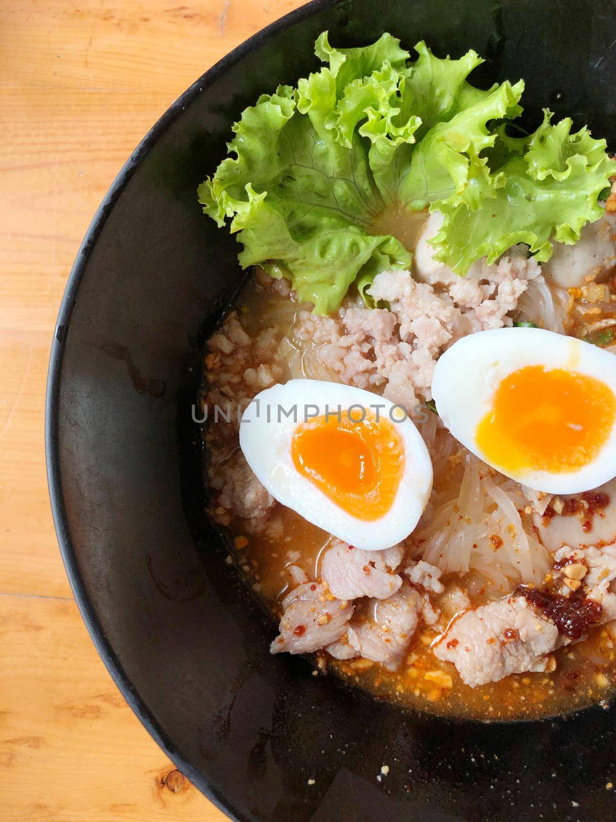 Thai spicy pork noodle soup with coconut milk, vegetables herbs and onsen tamago eggs  in black bowl with sesame seeds and peanut on wooden table, view from above