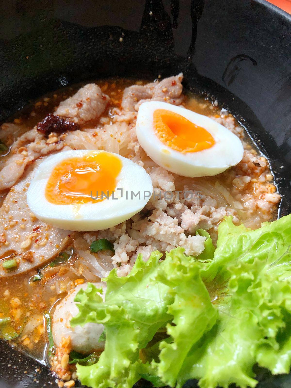 Thai spicy pork noodle soup with coconut milk, vegetables herbs and onsen tamago eggs  in black bowl with sesame seeds and peanut on wooden table, view from above by NarinNonthamand