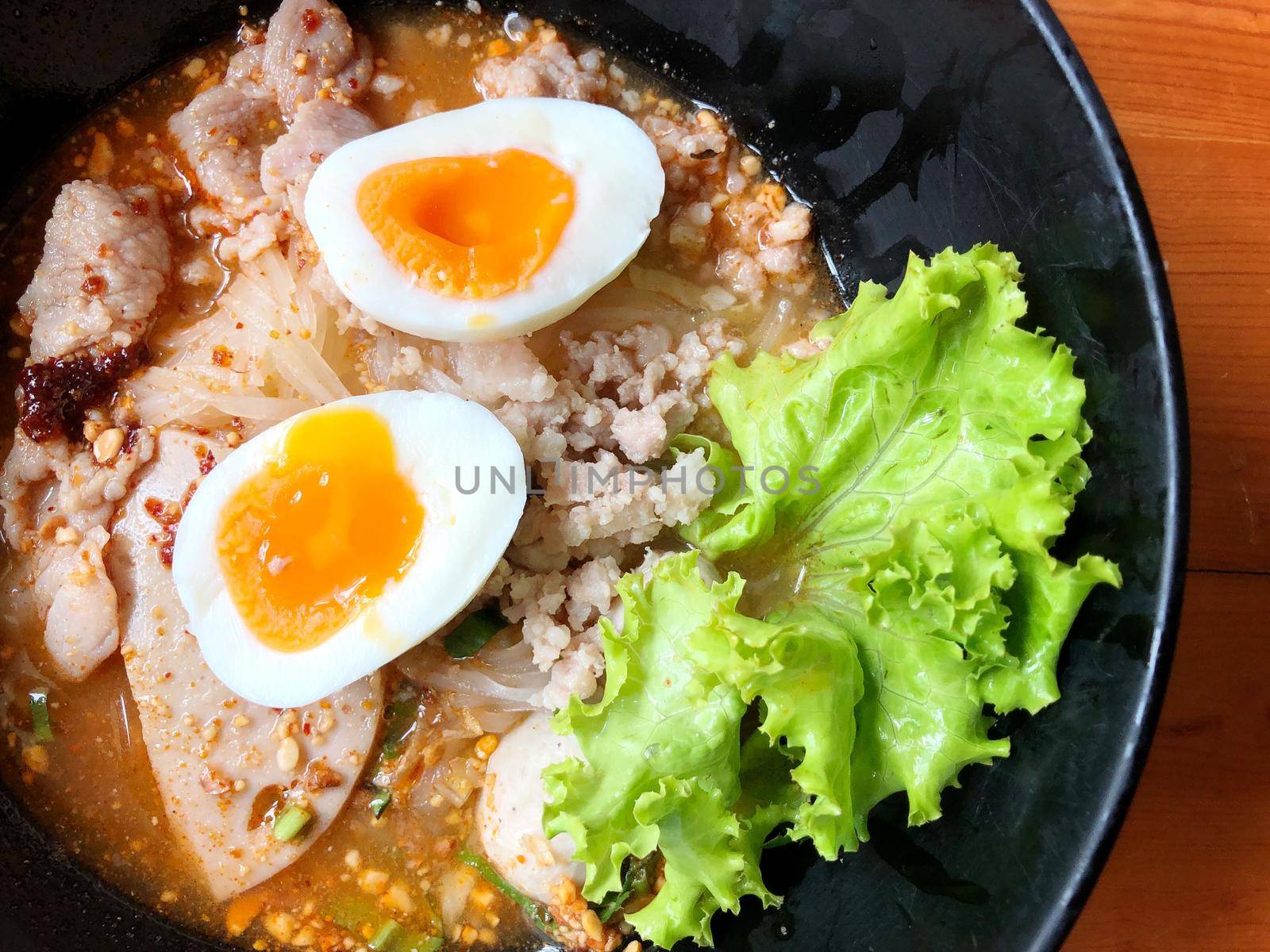 Thai spicy pork noodle soup with coconut milk, vegetables herbs and onsen tamago eggs  in black bowl with sesame seeds and peanut on wooden table, view from above by NarinNonthamand