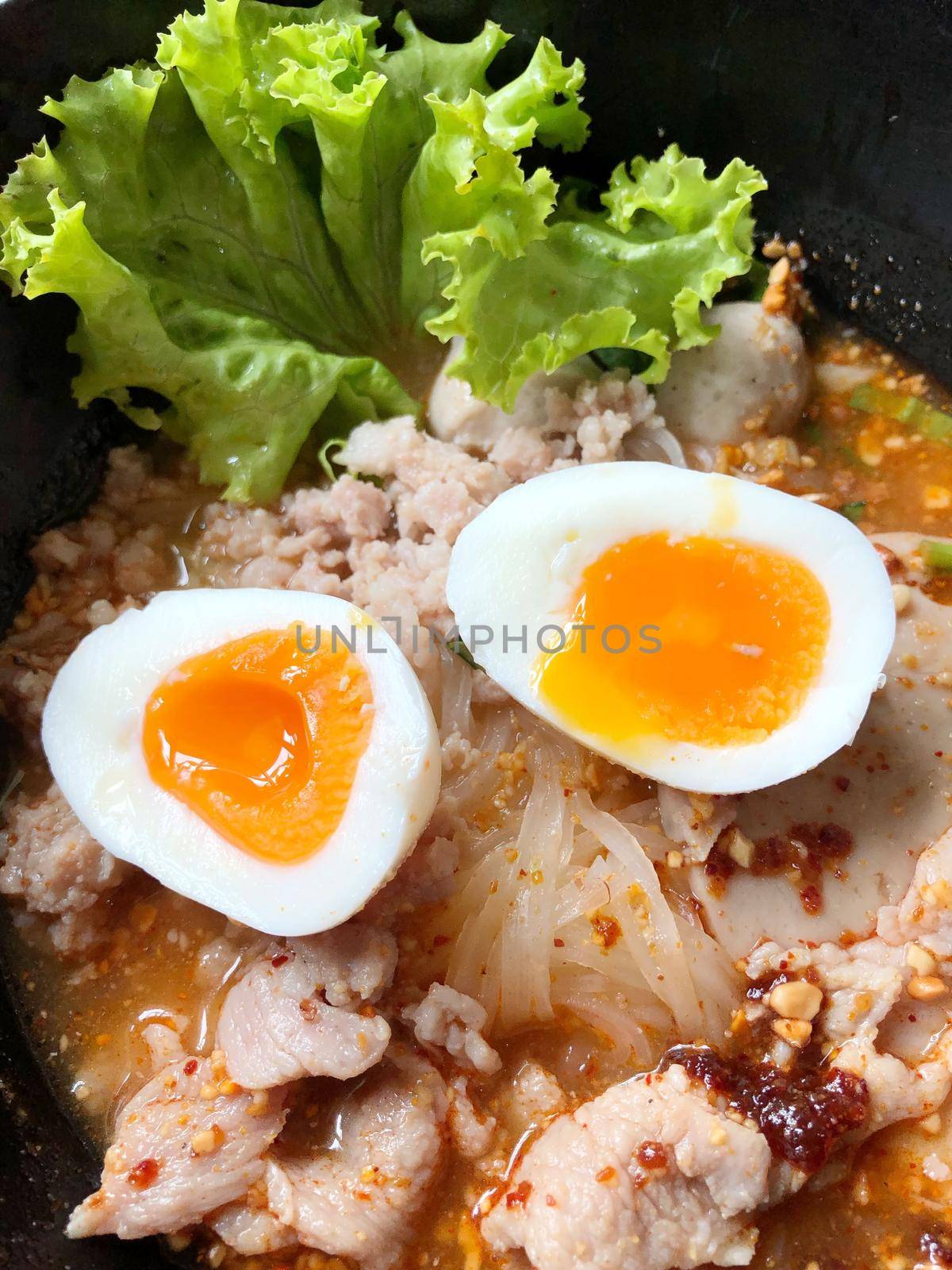 Thai spicy pork noodle soup with coconut milk, vegetables herbs and onsen tamago eggs  in black bowl with sesame seeds and peanut on wooden table, view from above