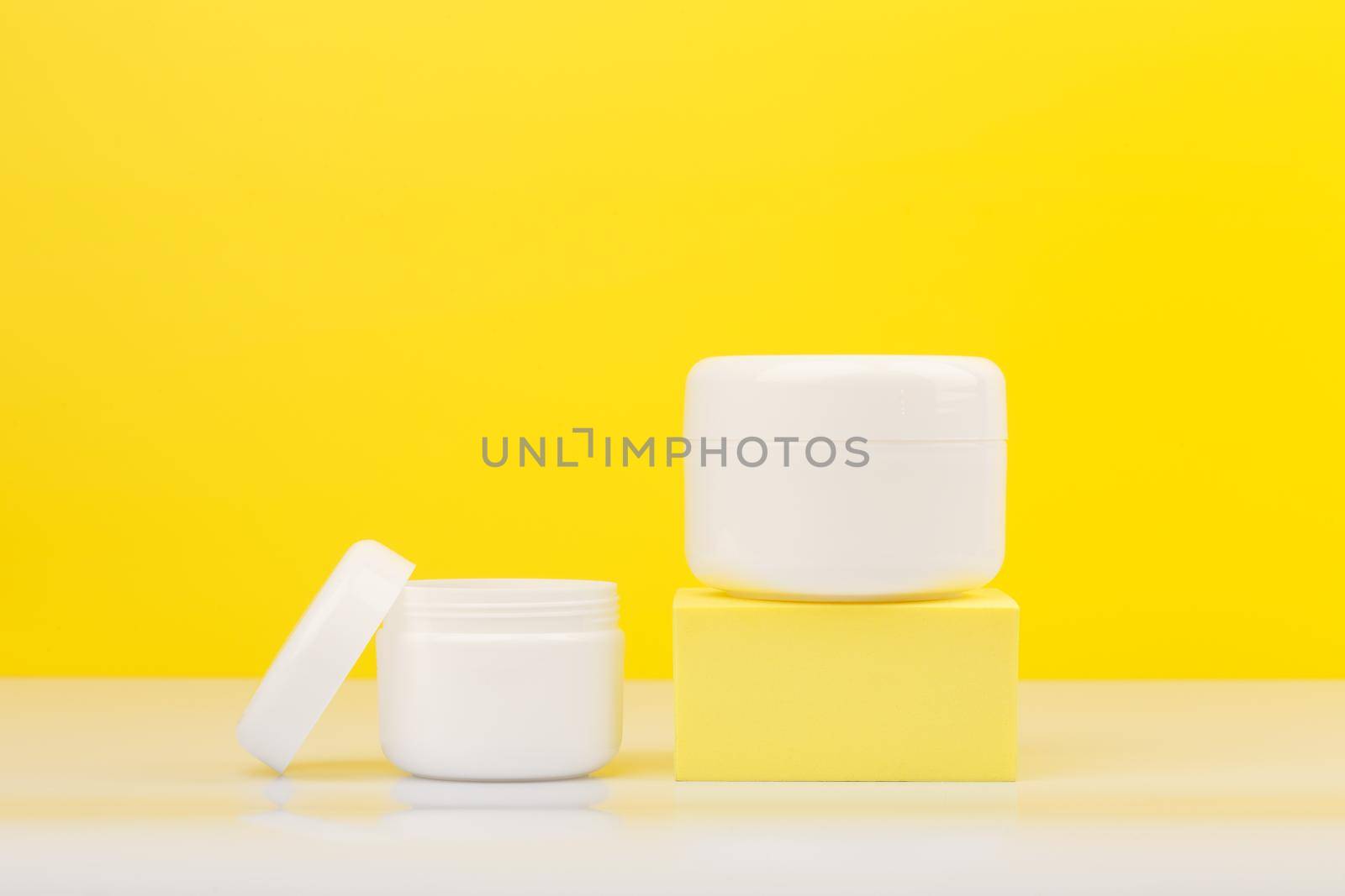 Two cream jar with the bigger one on podium on white glossy table against yellow background. Minimalistic concept of daily beauty routine and skin treatment