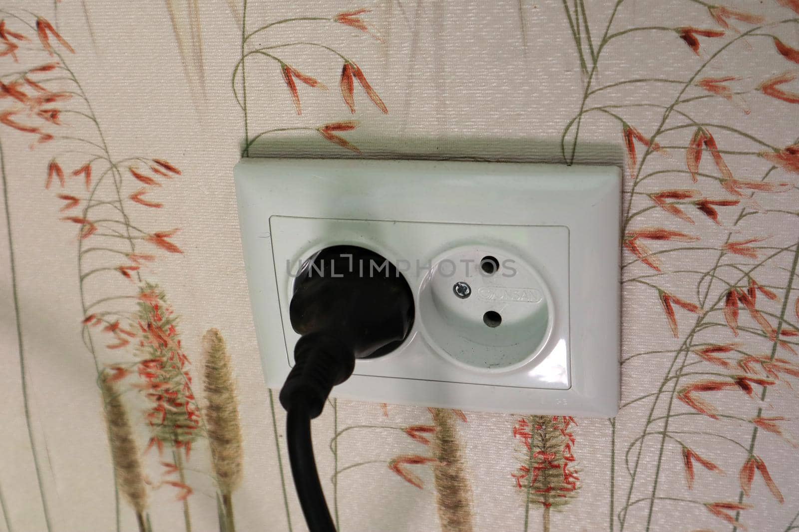 Double electrical power European socket with black cable and plug on wall with light wallpaper with a pattern of plants