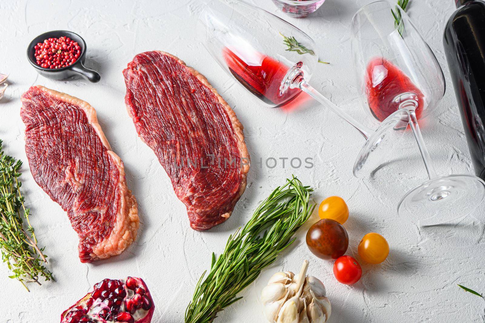 Picanha organic beef steaks with rosemary, peppercorns, pomegranate, near red wine in glasses and bottle over white textured background, side view.