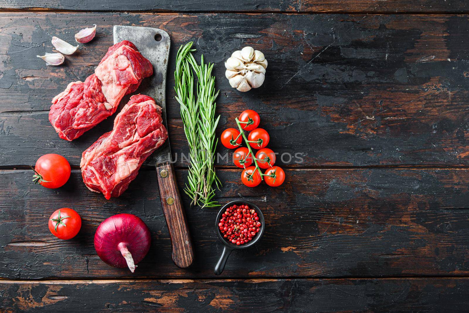 Organic raw chuck eye roll beef steak cut with rosemary red onin, garlic peppercorns and tomatoes on old dark wooden table. Top view with space for text
