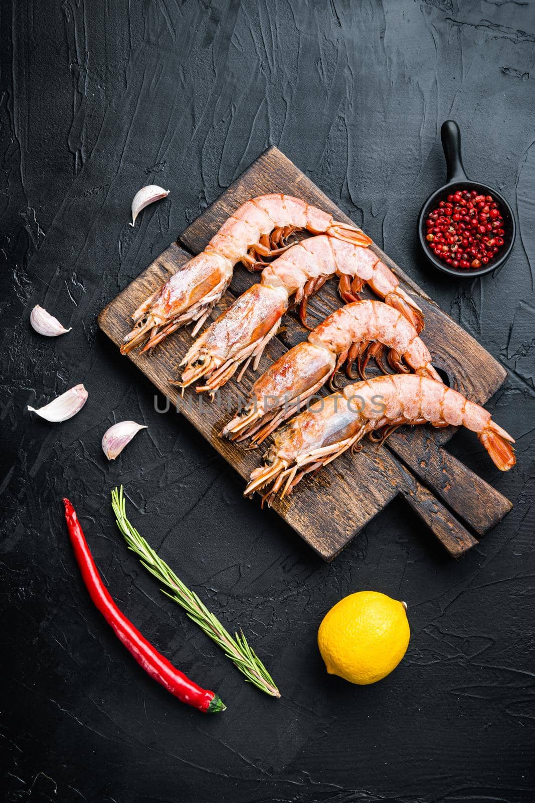 Seafood shrimps on wooden cutting board with spices, pepper and lemon on black concrete surface, flat lay, food photo.