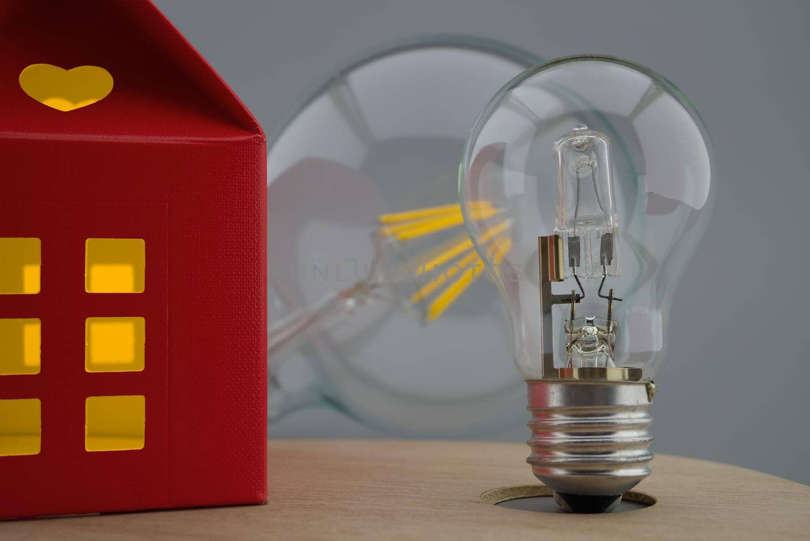 Incandescent lamp light bulbs next to a small red house of paper with a cut out heart and warm light in windows. Close-up concept of home lighting