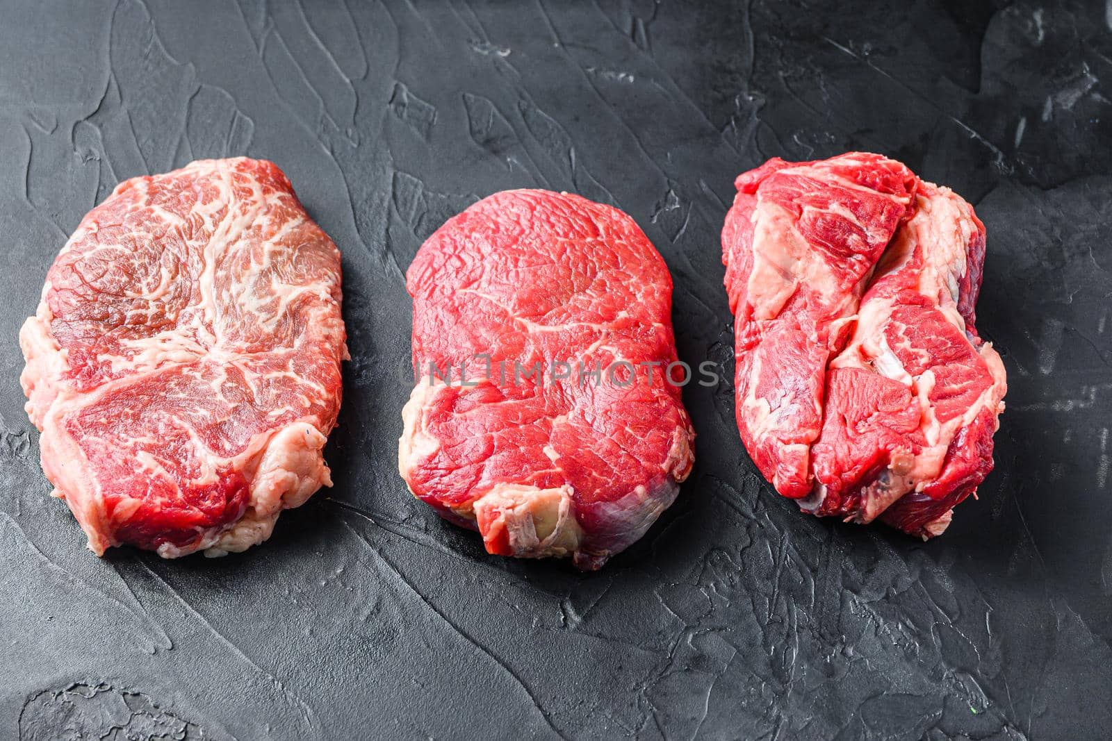 Set of raw alternative cuts beef steaks, top blade, rump and chuck roll, on black textured background, side view