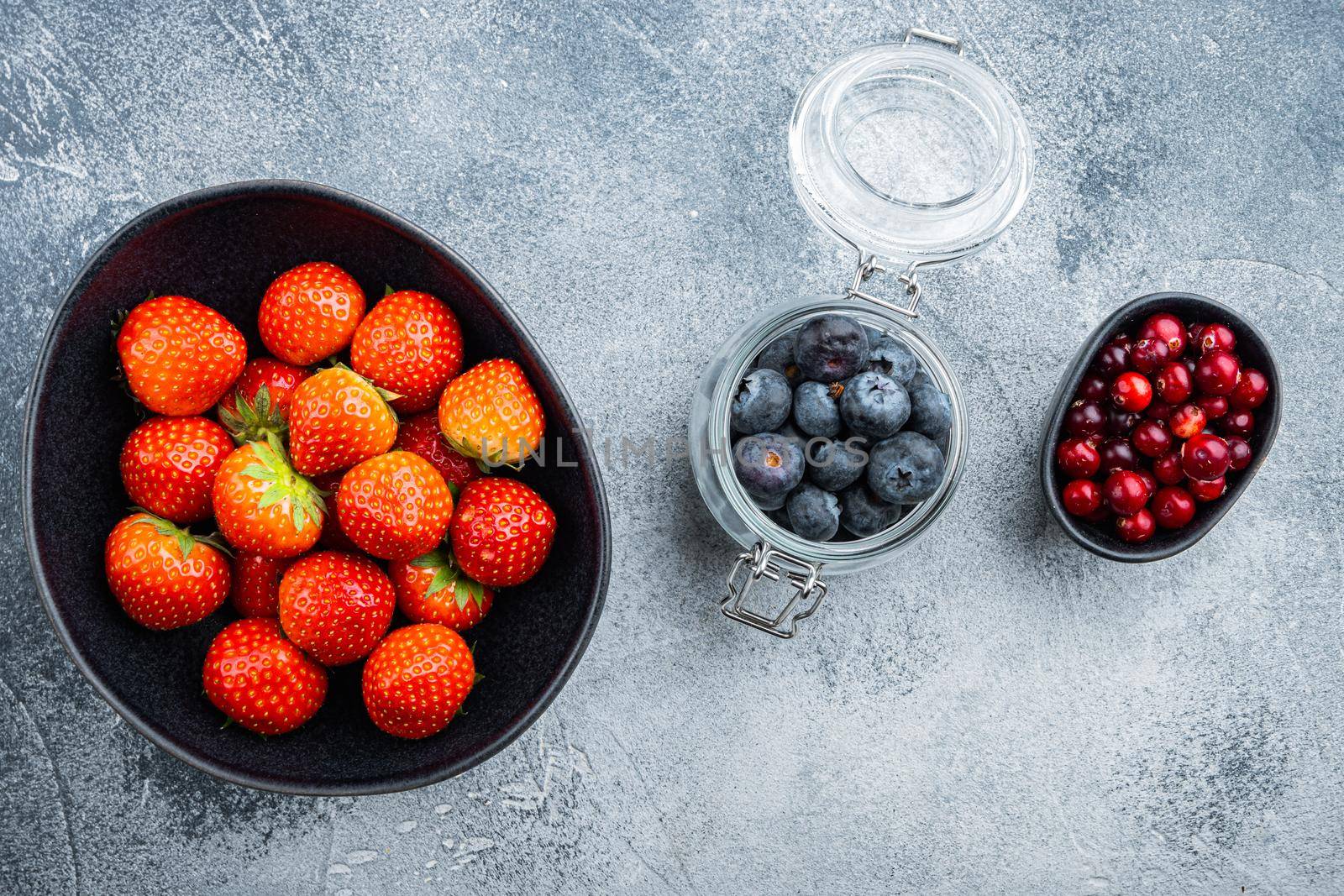 Ripe strawberry, cranberry and blueberry in bowl and jar, top view, on textured background.