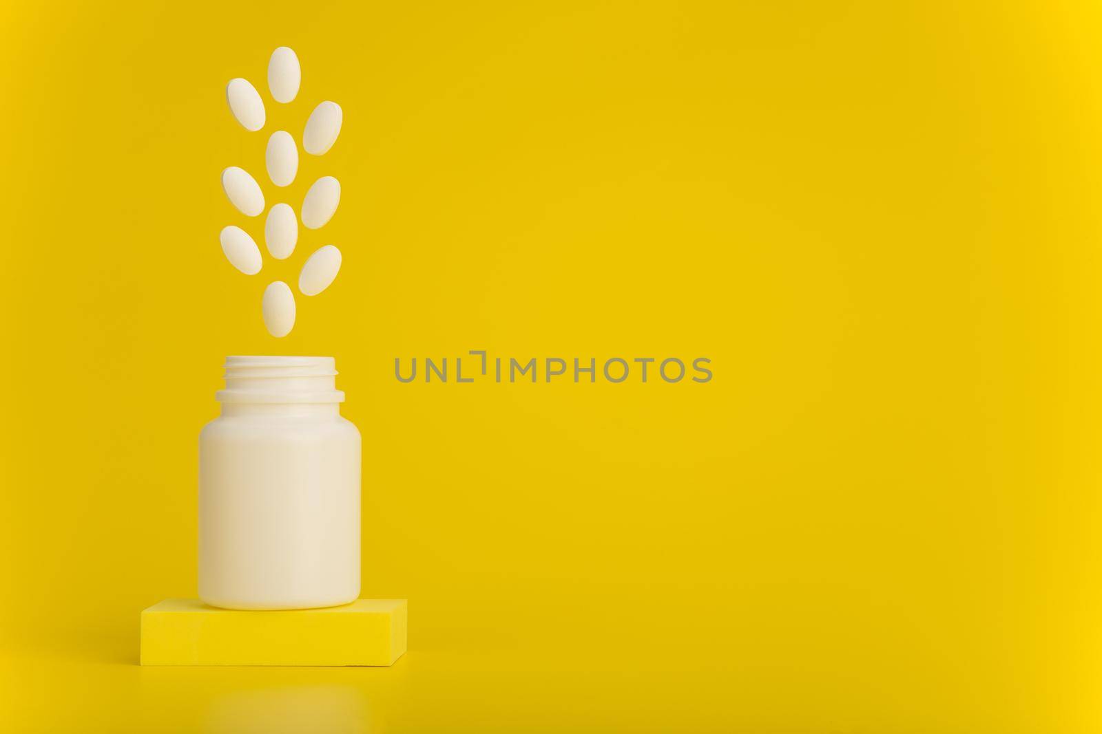 Still life with opened medication bottle on podium with white oval pills jumping out against yellow background with copy space. Concept of vitamins for kids or wellbeing and pharmacy
