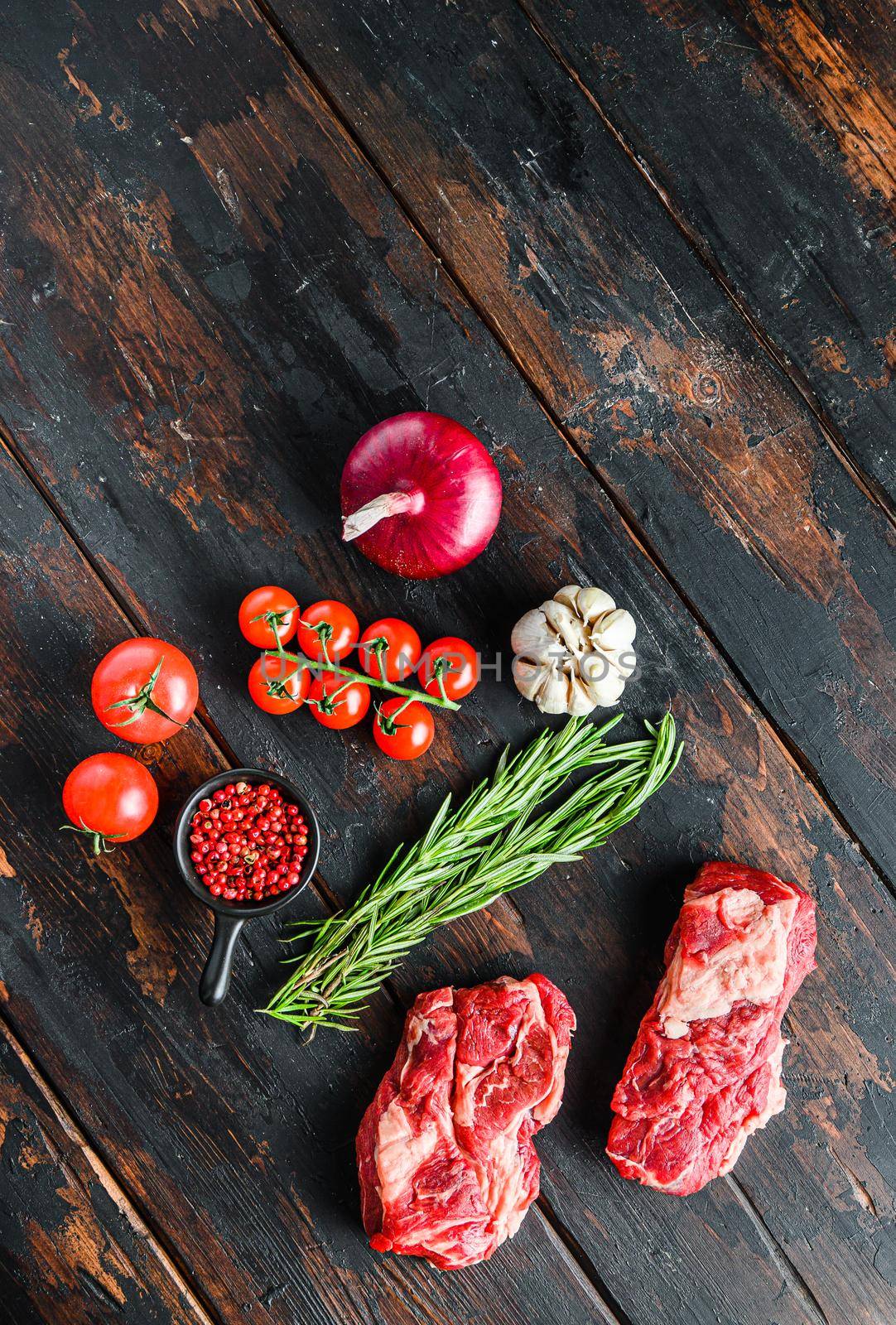 Raw chuck eye roll beef steak cut with rosemary red onin, garlic peppercorns and tomatoes on old dark wooden table. Top view with space for text