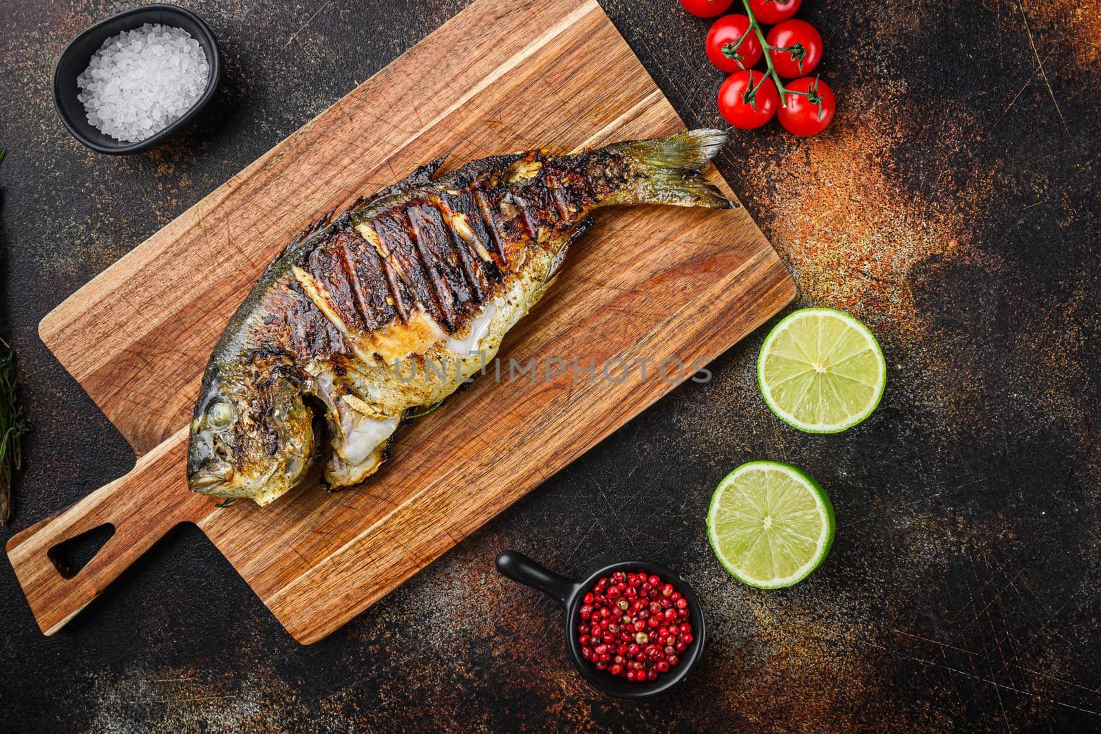 Grilled sea bream or dorado raw fish on chopping board with ingredients over old metall dark background, top view with space for text