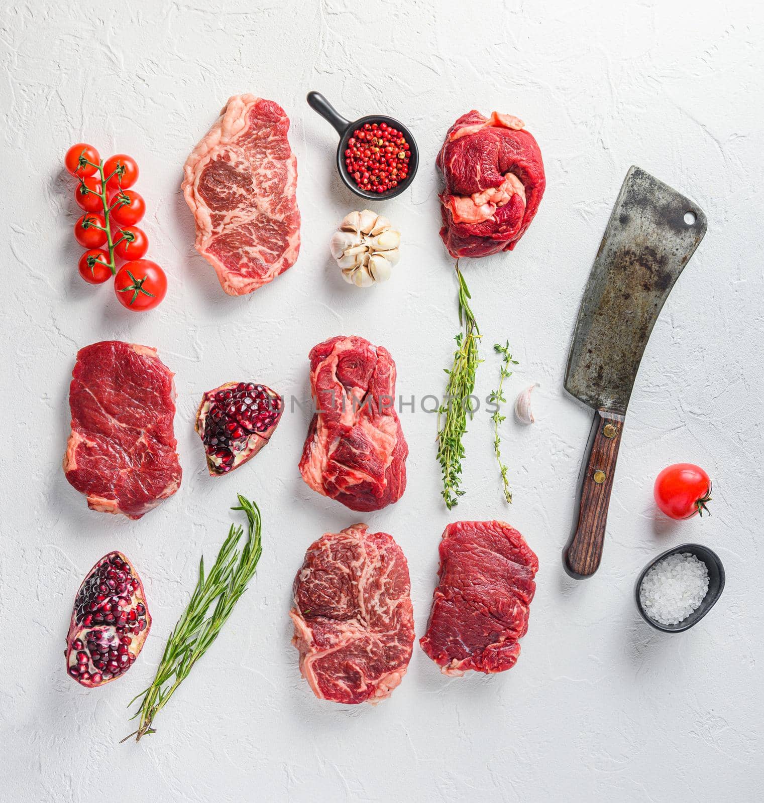 Rraw beef steaks set top blade, rump, chuck eye roll with herbs pomegranate and butcher knife for chopping over white concrete background, top view
