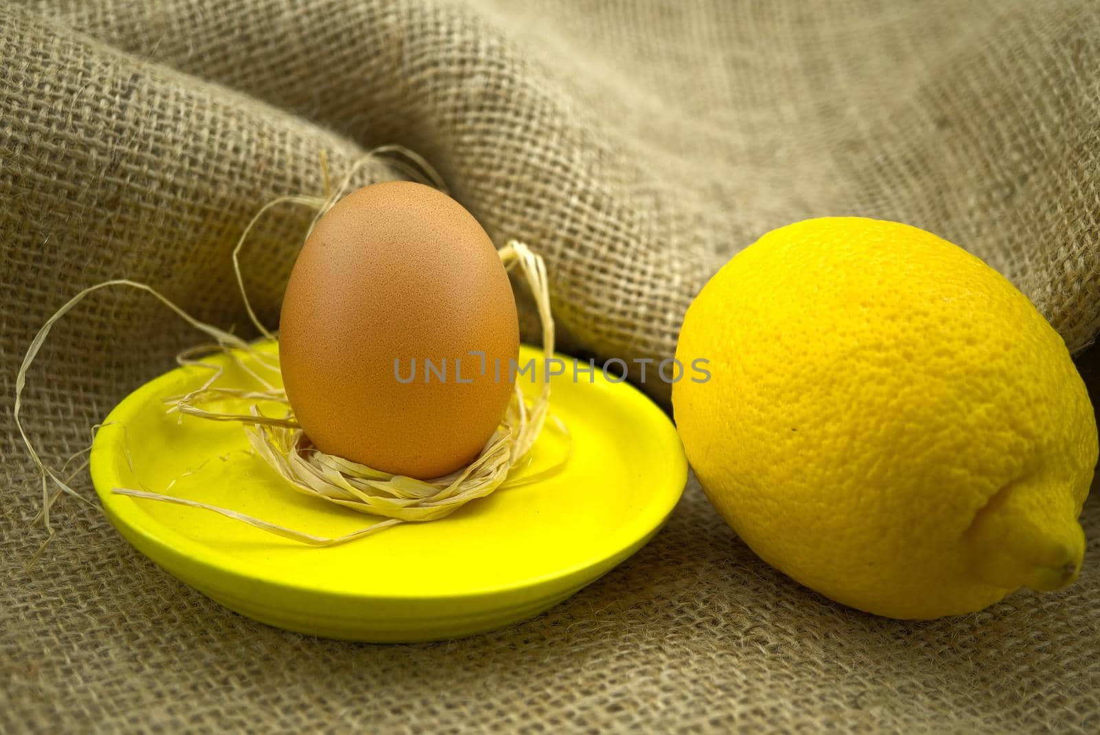 Brown egg on bright yellow plate near yellow lemon over rustic hessian sack background. Organic farming and color trends concept with copy space