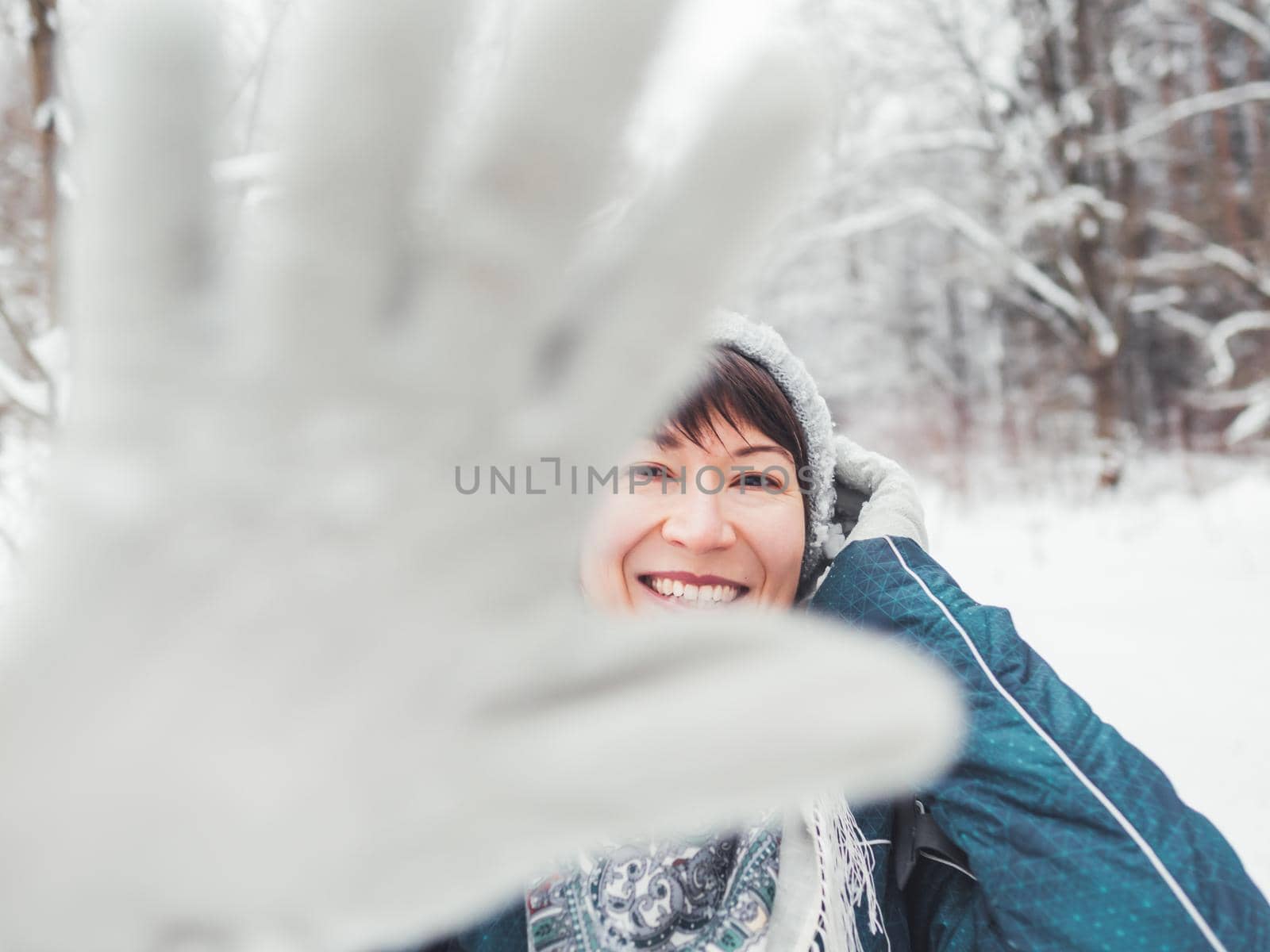 Smiling woman is closing camera with hand in warm glove. Fun in snowy winter forest. Woman laughs as she walks through wood. Sincere emotions.