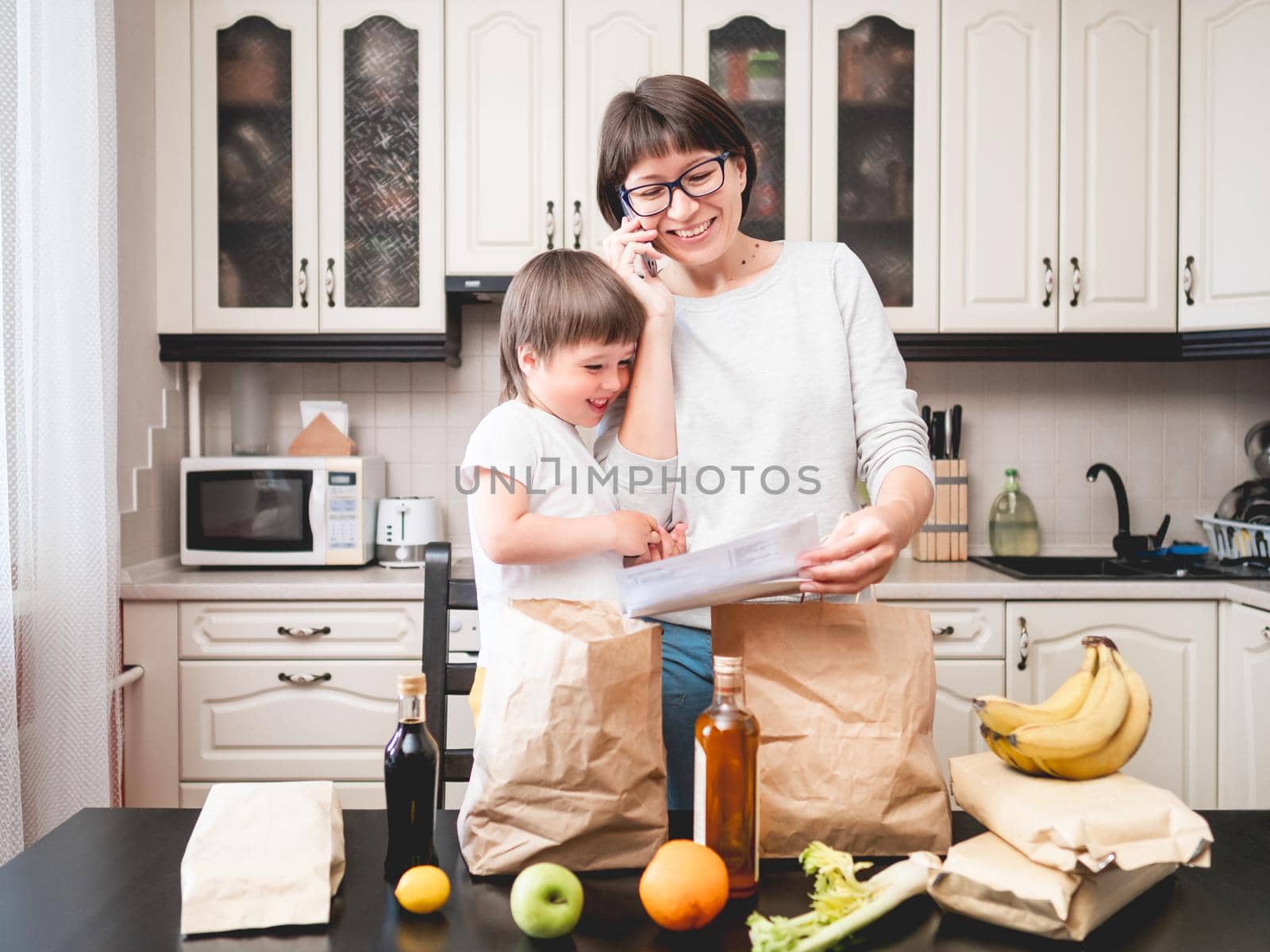 Woman and toddler boys sorts out purchases in the kitchen. Grocery delivery in paper bags. Online order from grocery store. Mother and son at kitchen. by aksenovko