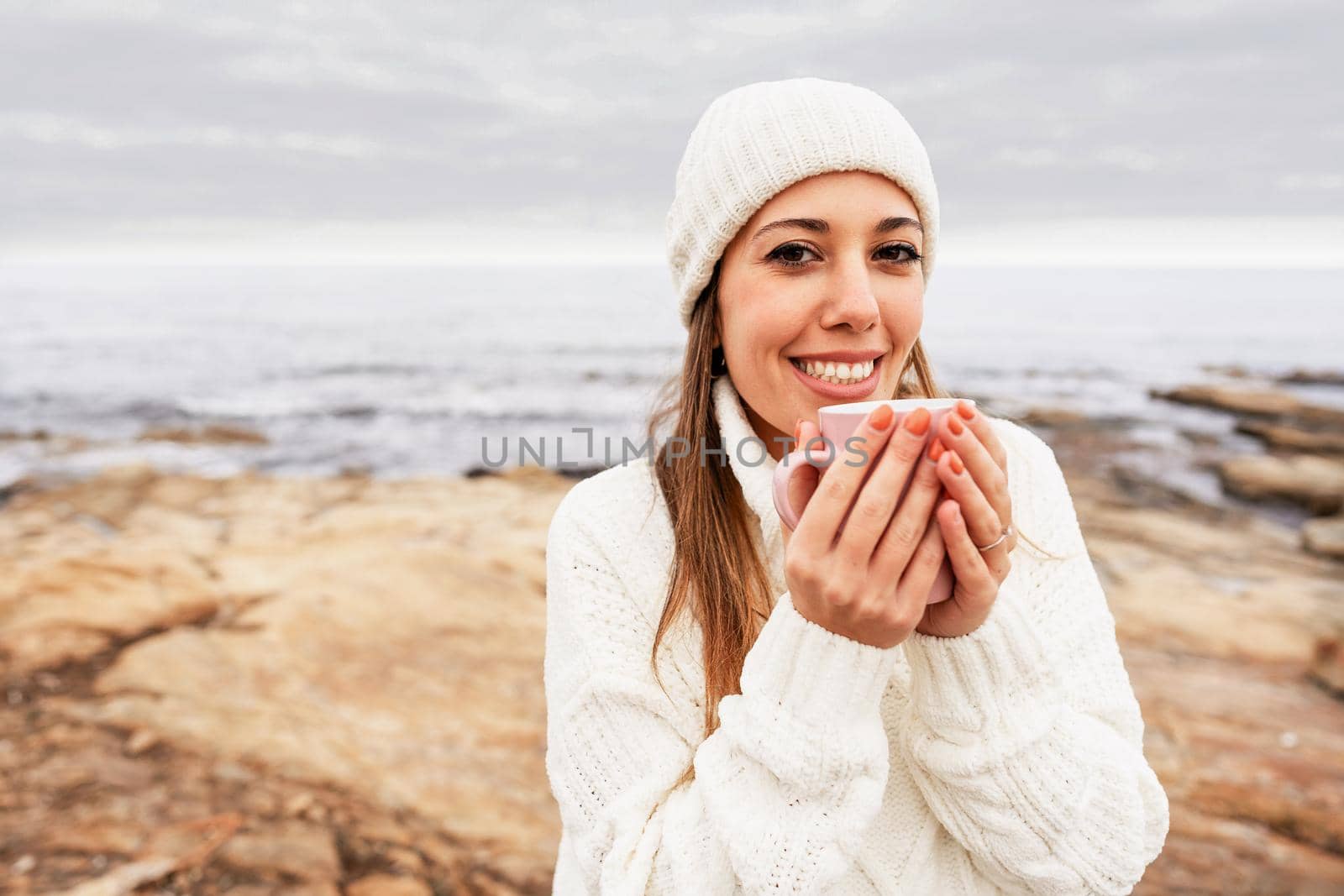 Beautiful young caucasian confident woman looking at camera with toothy smile wearing white wool hat and sweater holding a cup in his hands outdoor at a natural sea resort in winter vacation