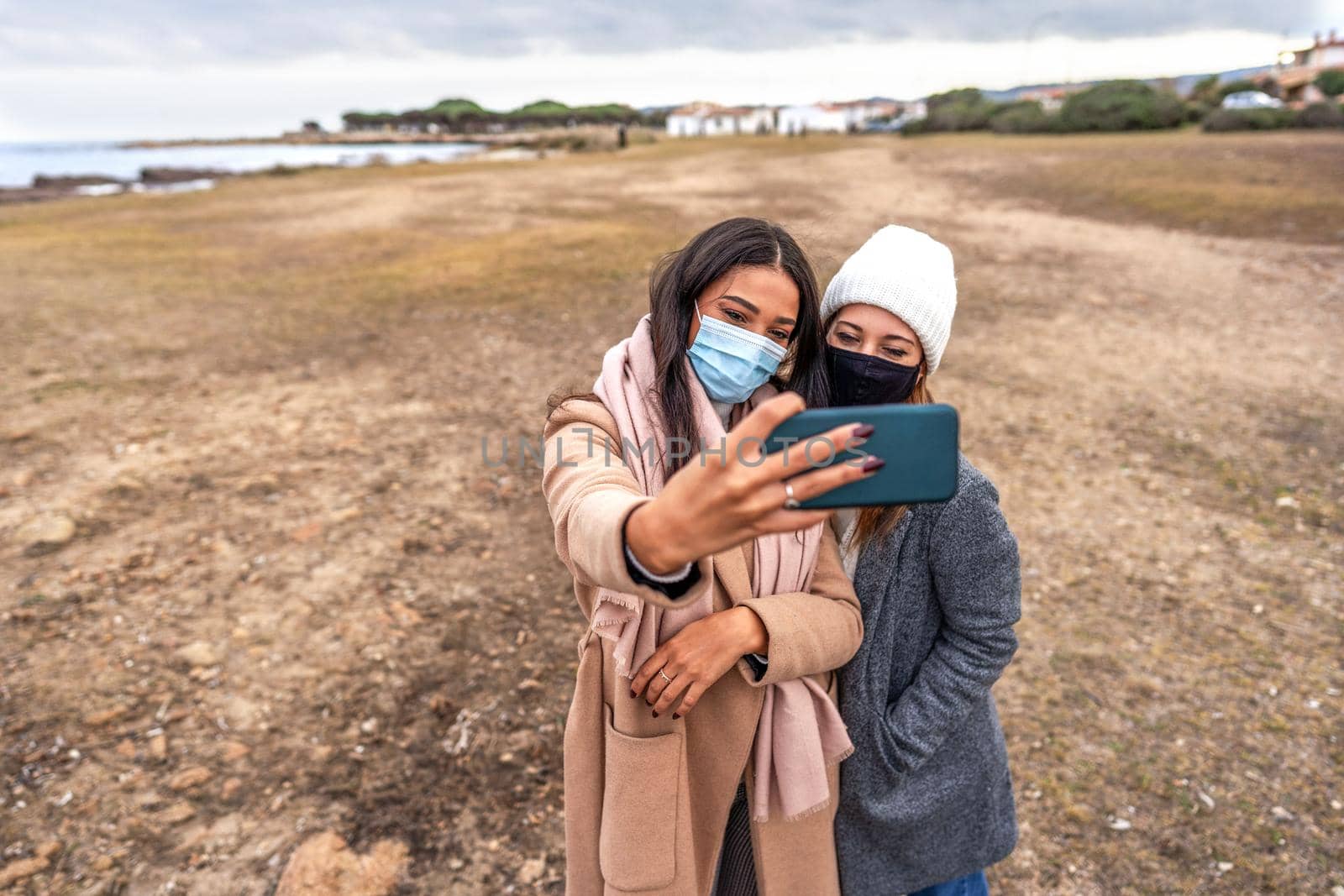 Two happy girls having fun outdoor on vacation at sea resort doing selfie with smartphone wearing protective mask. Multiracial female couple in winter clothes using technology to make a self-portrait