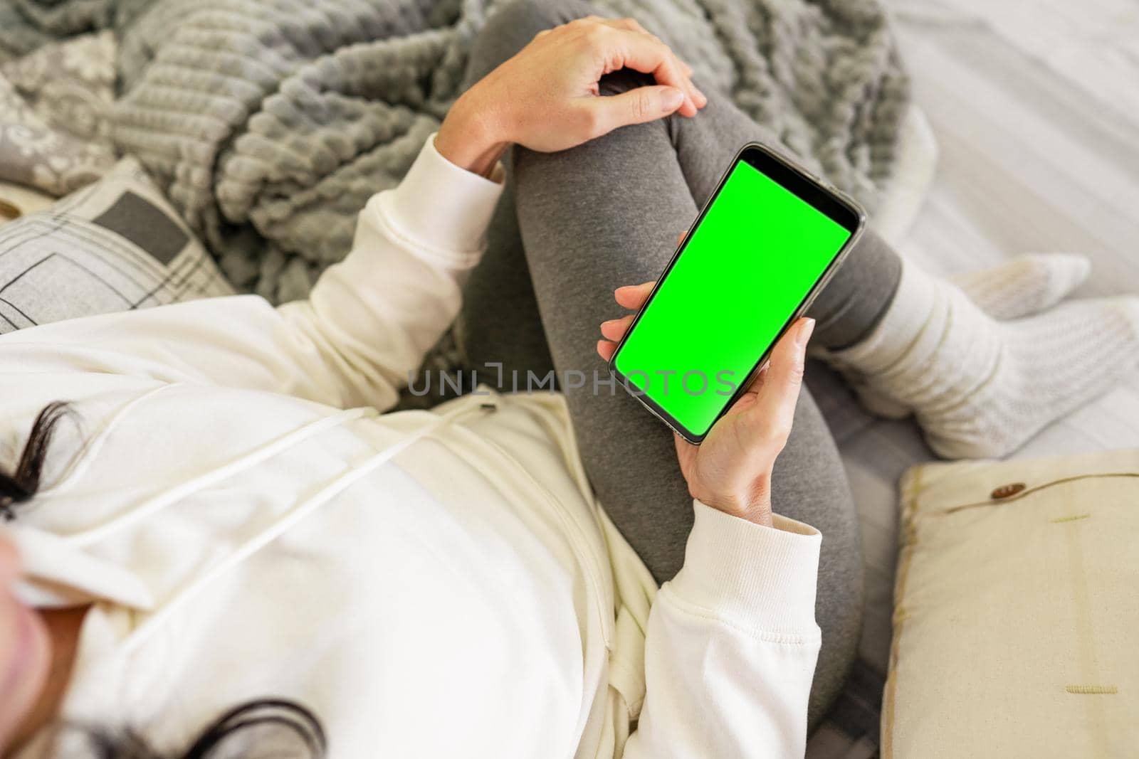 Top view of unrecognizable young woman on sofa holding smartphone with green screen blank display for copy space. Caucasian female beautiful housewife relaxing in living room using internet mobile by robbyfontanesi