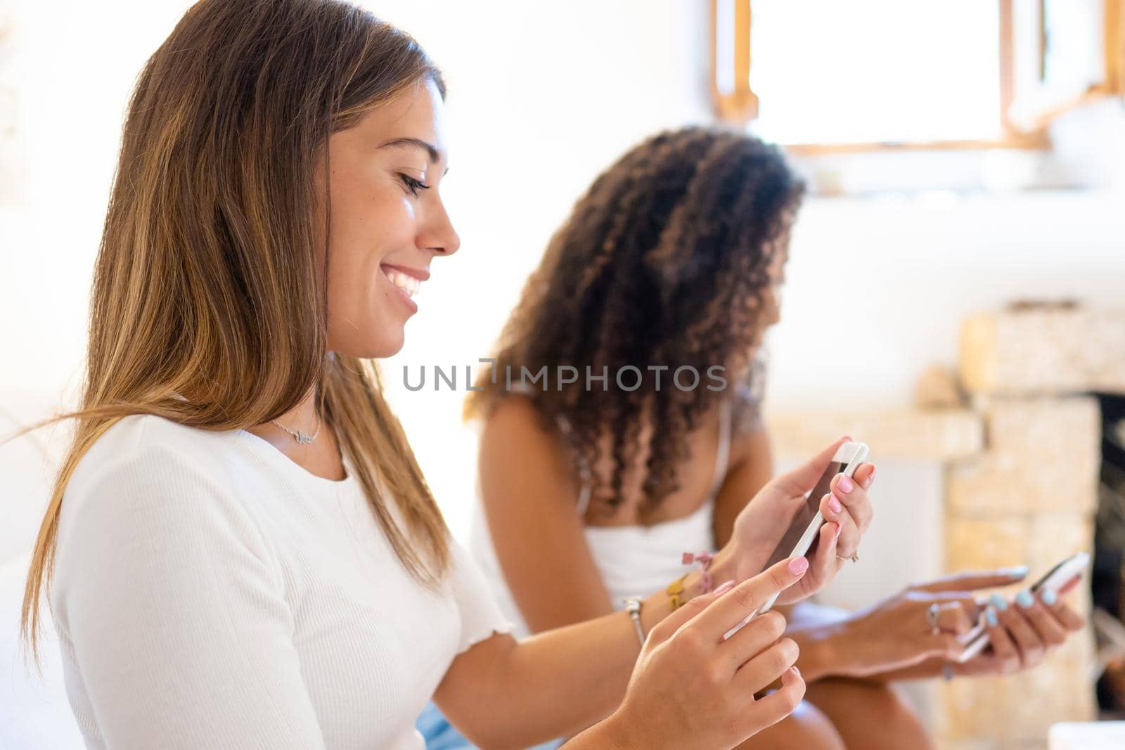 Bright vivid color shot of two young beautiful women sitting on sofa at home using smartphone. Selective focus close up of Caucasian girl chatting on social network with her female best friend blurred