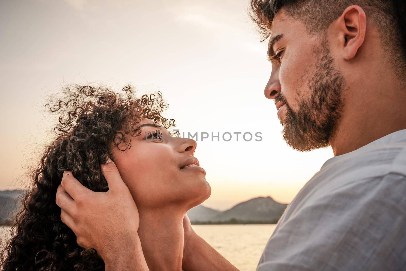 Close up portrait of young mixed race couple in love looking in eyes each other in sunset color mood. Macho dominant handsome man holding face of his worshiping submissive woman with intense gaze by robbyfontanesi