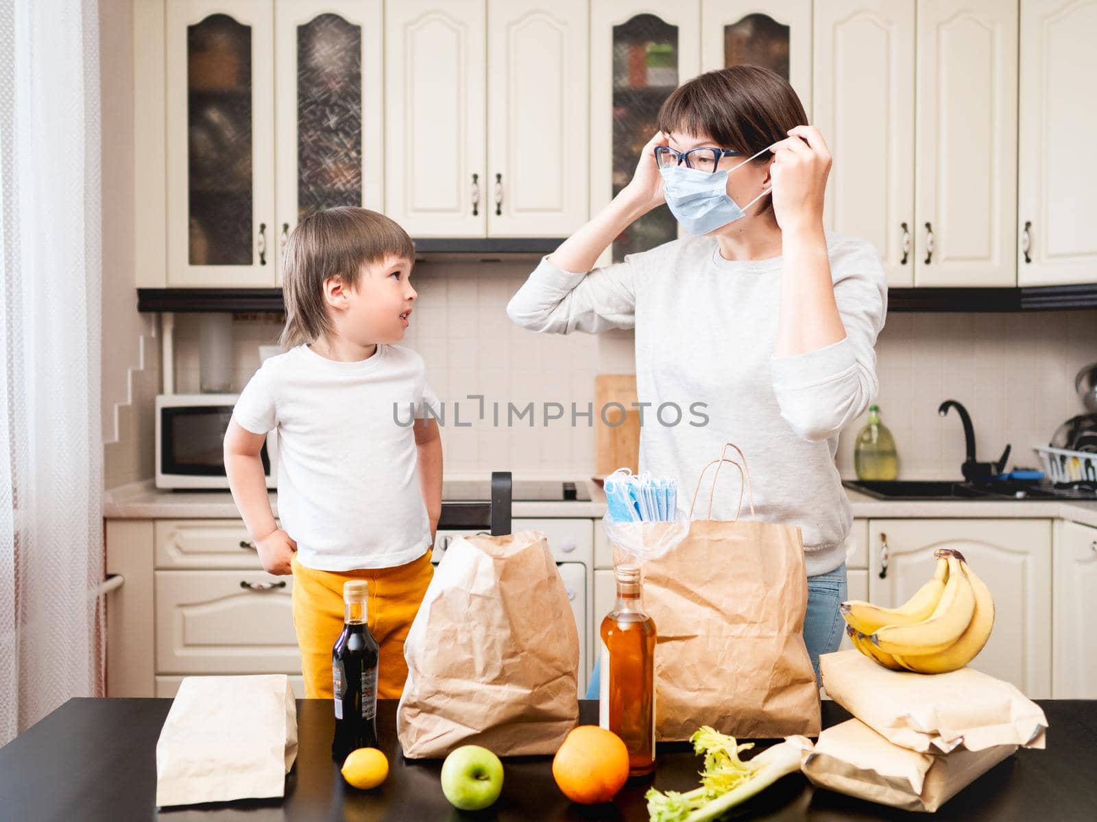 Woman and toddler sorts out purchases. Mom shows how to wear medical protective mask. Grocery delivery in paper bags. Online order from grocery store. Mother and son at kitchen. by aksenovko