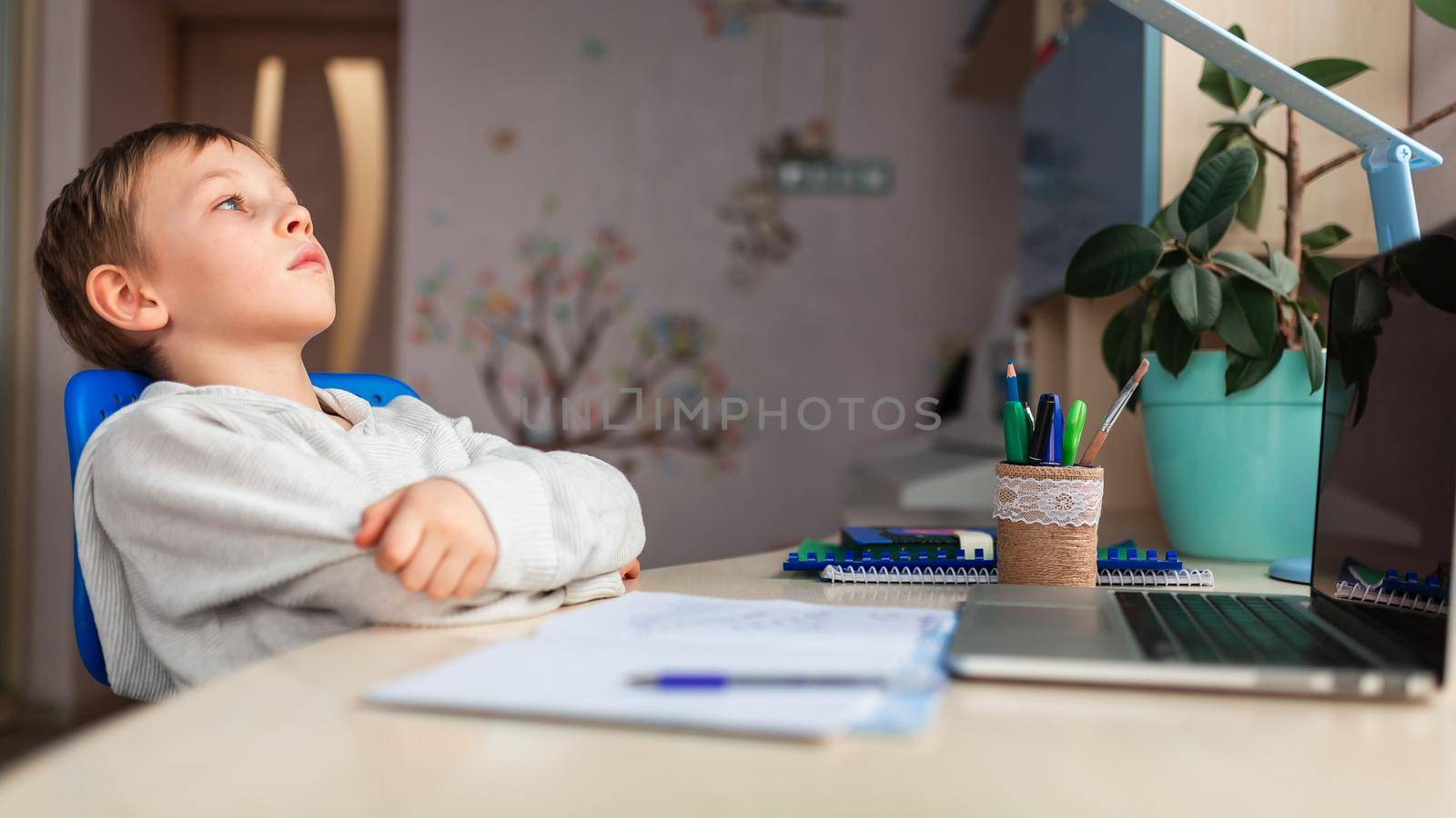 Cute little tired schoolboy studying at home doing school homework. Training books and notebook on the table. Distance learning online education