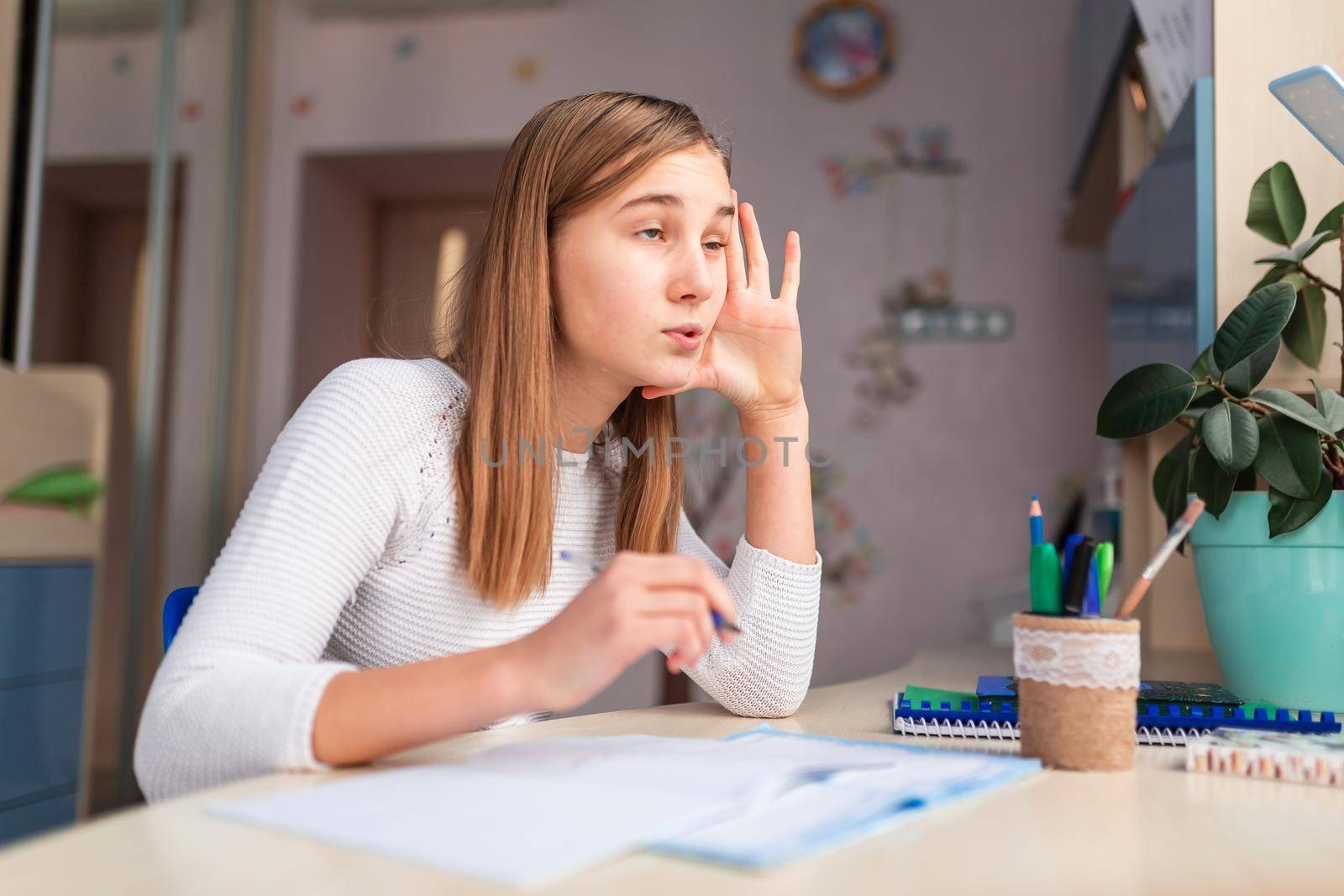 Beautiful bored schoolgirl studying at home doing school homework. Training books and notebook on the table. Distance learning online education