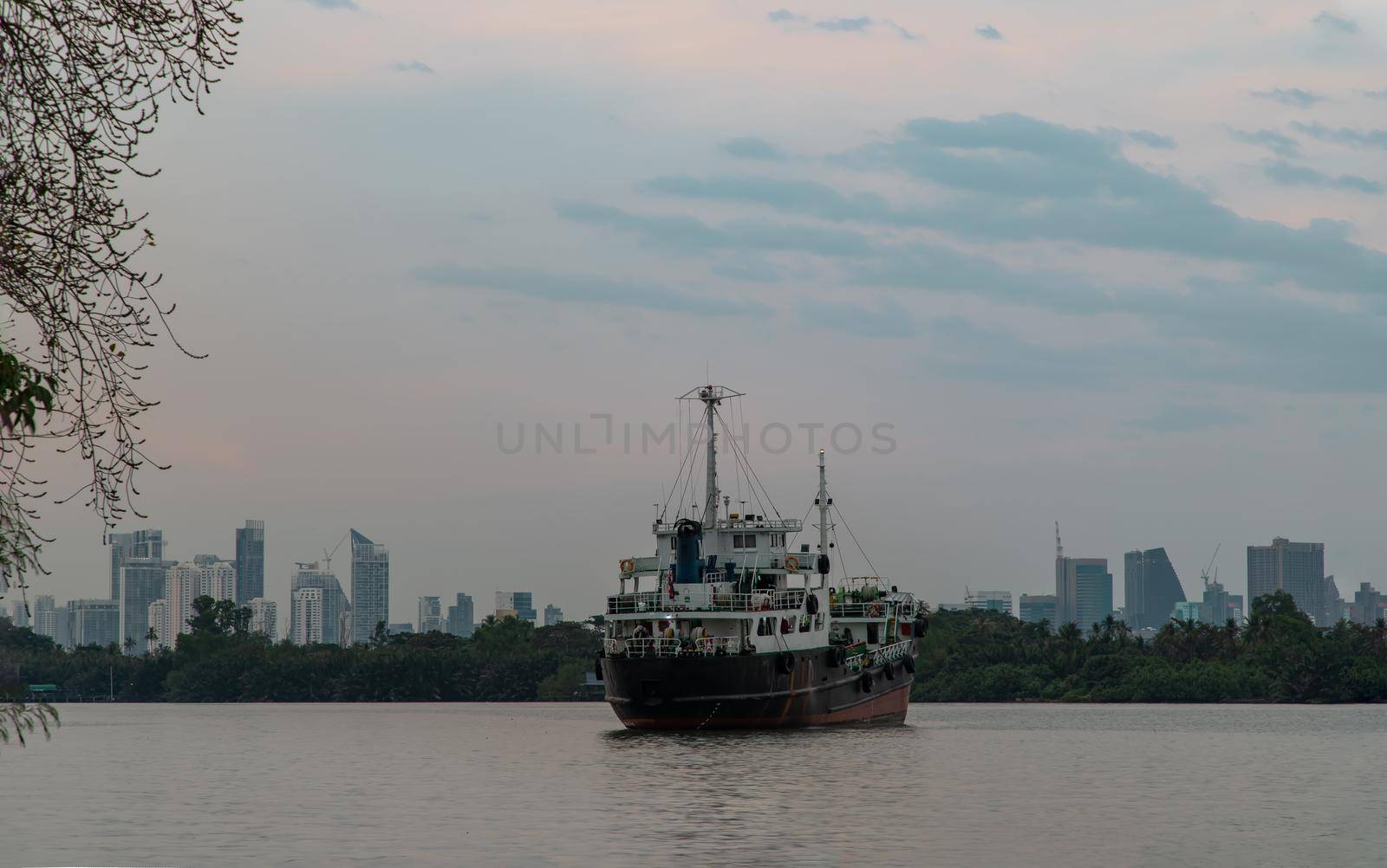 Bangkok, Thailand - 04 Jan 2021 : A cargo ship parked in the middle of the Chao Phraya River at evening with green forest and skyscrapers at the background. Selective focus.